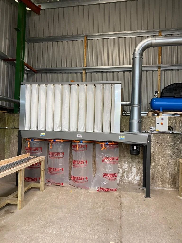 Industrial dust collector system with white filter bags, metal duct, and supports in a workshop or warehouse.