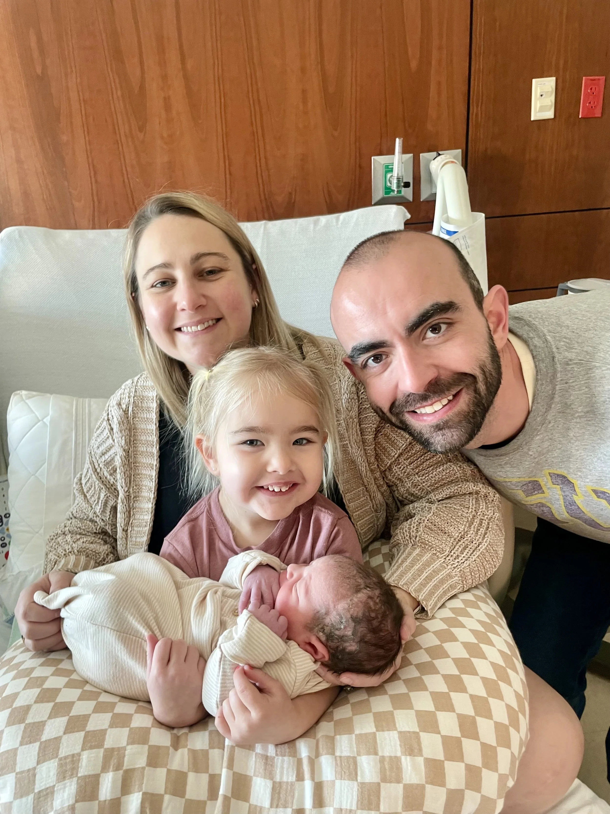 A family of four with two young children, including a newborn, gathered on a bed in a hospital room, smiling at the camera.