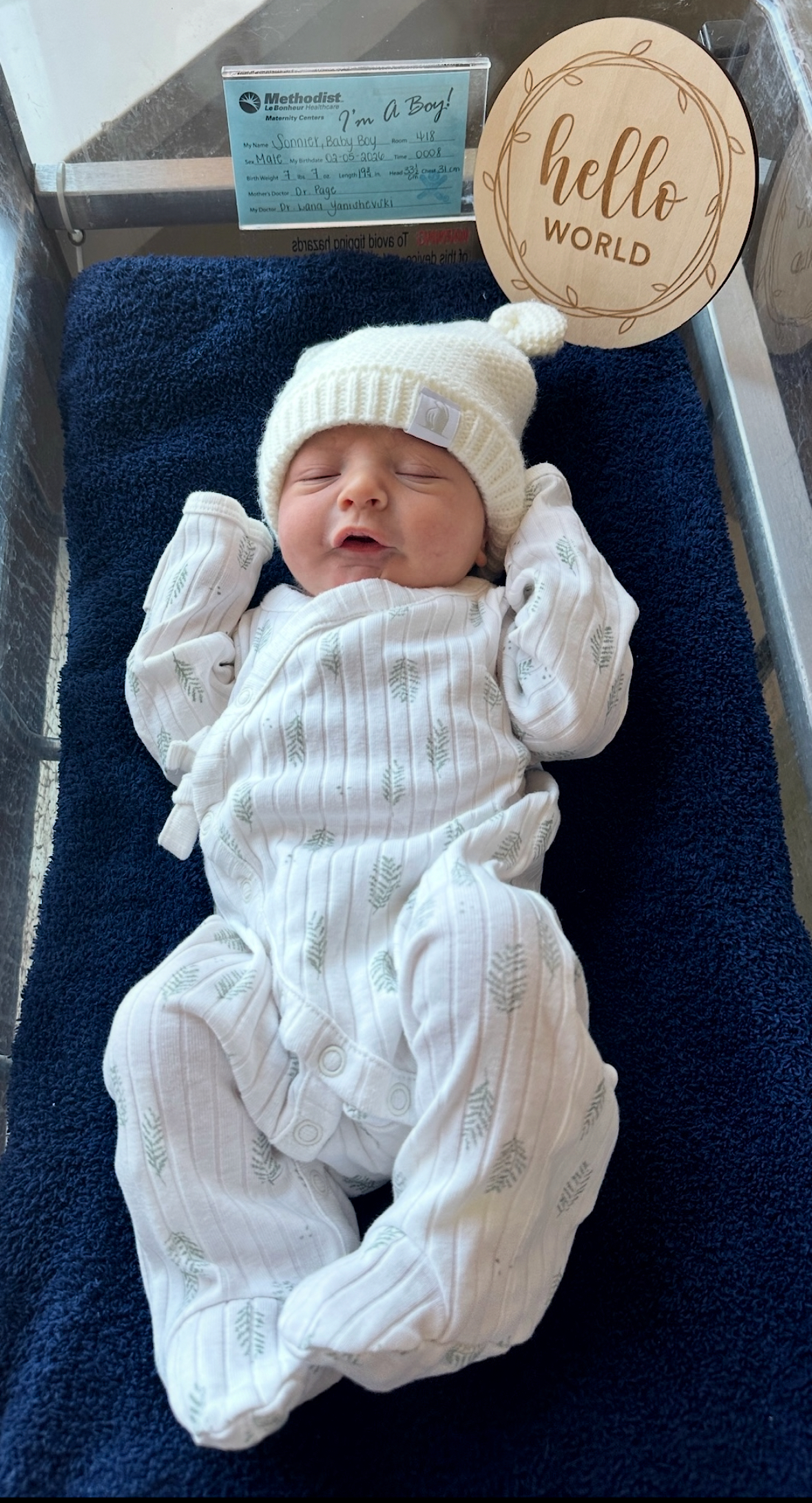 A newborn baby in hospital bassinet, wearing a white hat and onesie with green leaf pattern, sleeping with lips pursed. There is a sign that says 'I'm a Boy!' and a round wooden sign with 'hello WORLD' written on it.