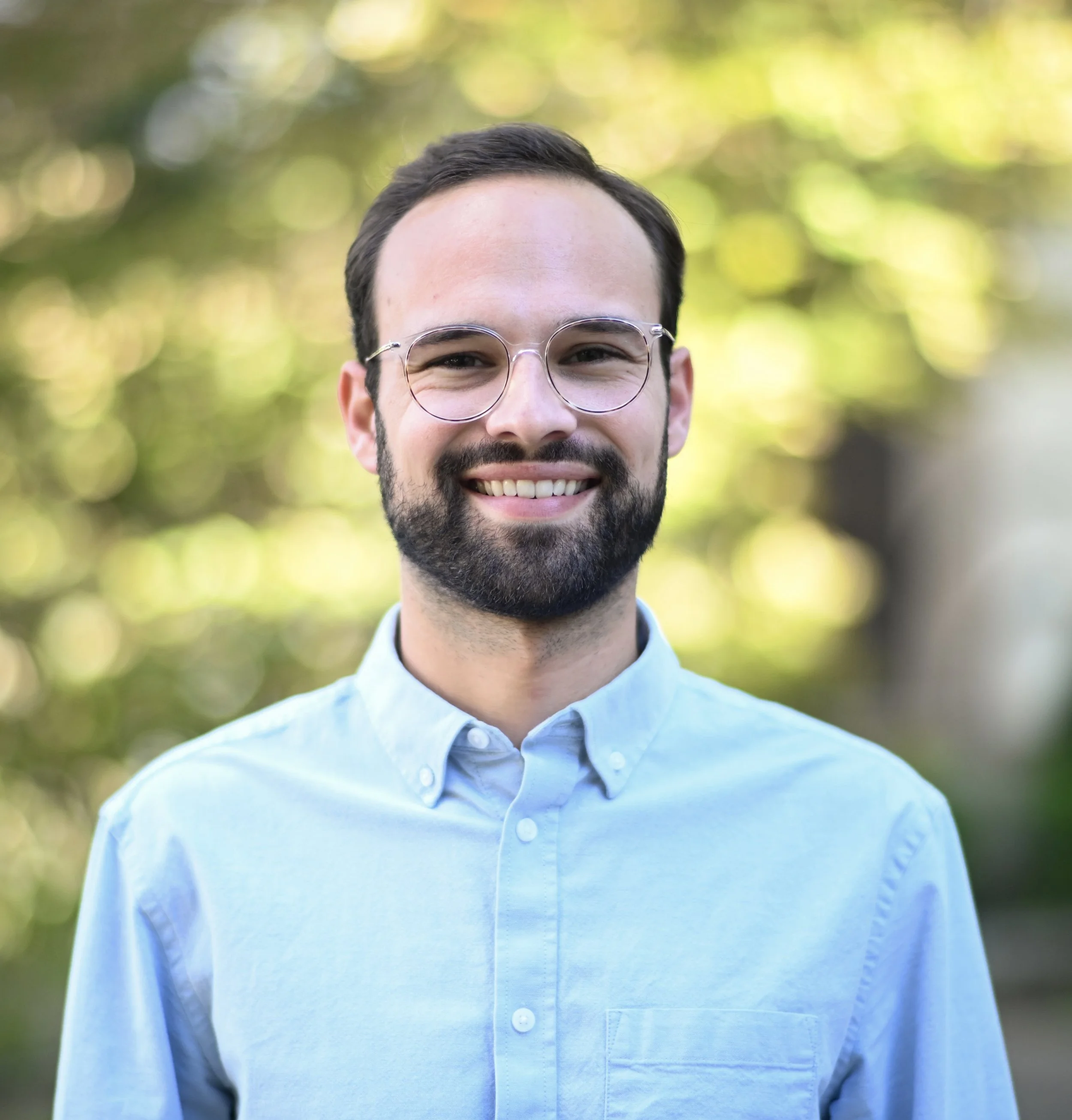 Close-up of a smiling young man with dark hair, beard, and glasses wearing a light blue button-up shirt, standing outdoors with blurred green and yellow foliage in the background.
