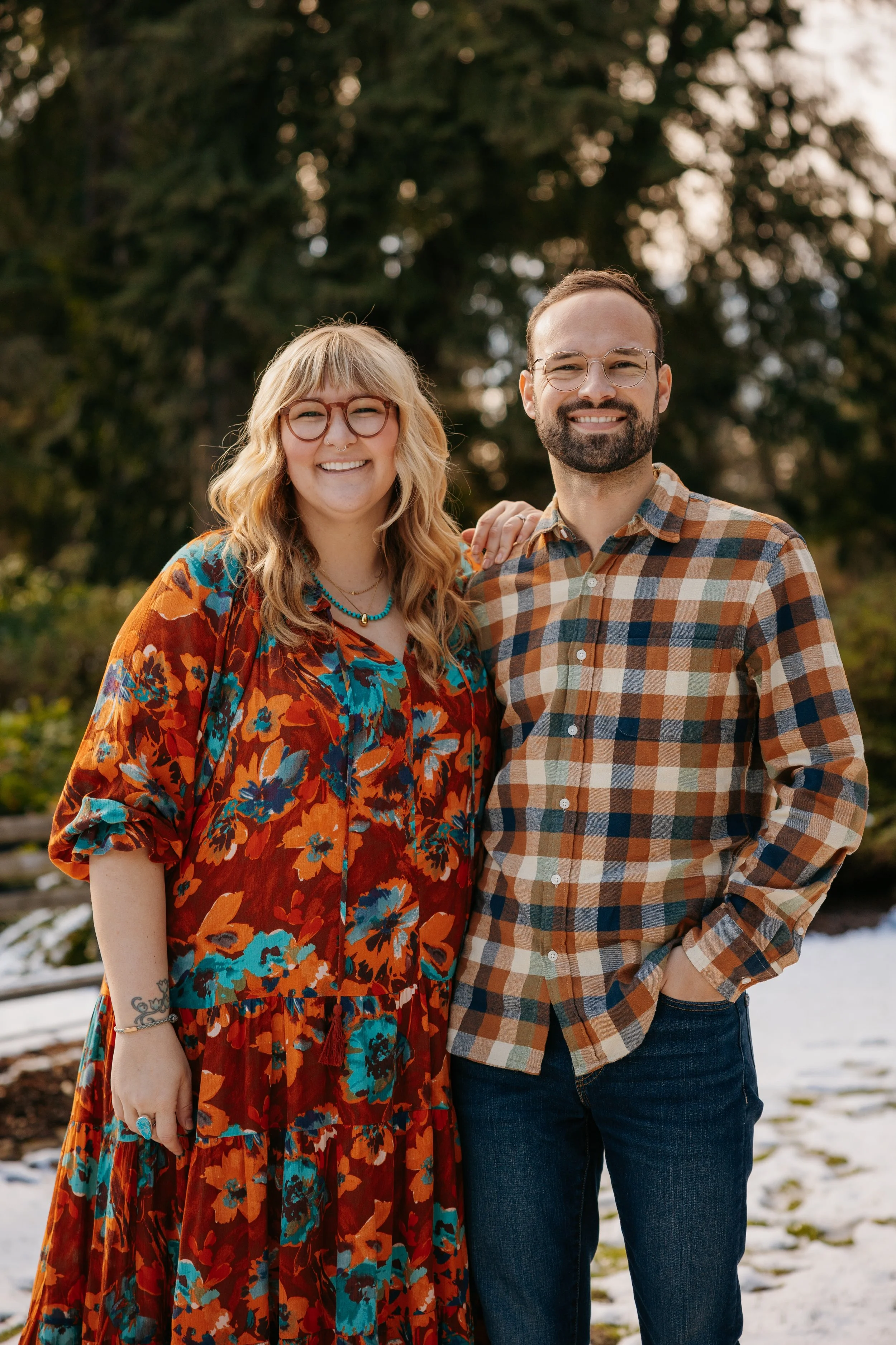A smiling woman with blonde hair, glasses, and a floral dress standing next to a smiling man with a beard, glasses, and a plaid shirt outdoors in a park with trees and snow on the ground.