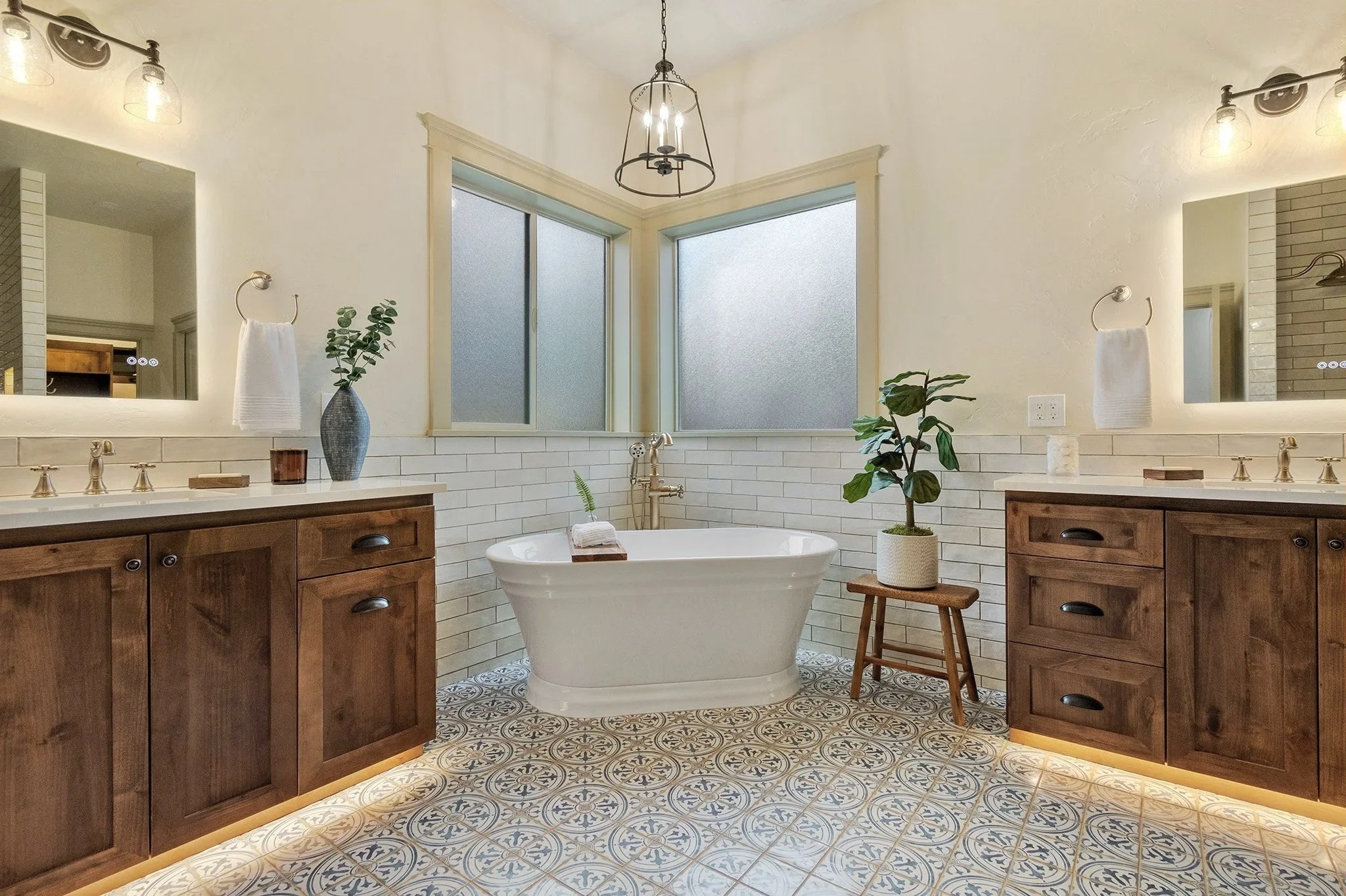 A bathroom with a freestanding bathtub in front of large frosted windows, with wood vanity cabinets on either side, plants, mirrors, and patterned floor tiles.