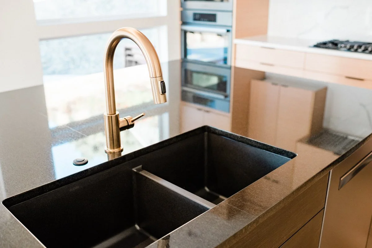 Modern kitchen with black double sink and gold faucet, surrounding gray countertop, and built-in appliances in the background.