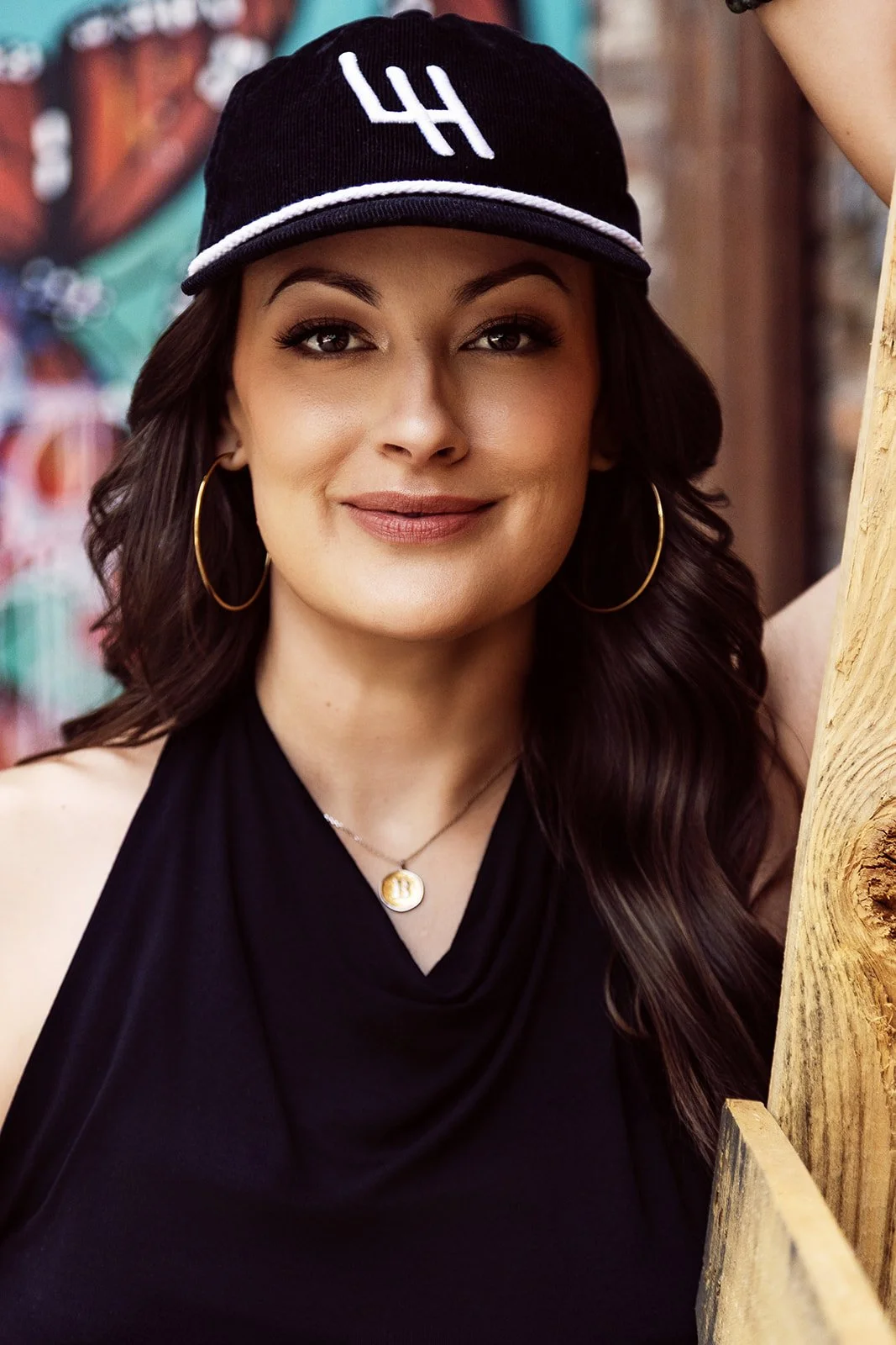 A woman with long dark hair, wearing hoop earrings, a black sleeveless top, a gold pendant necklace, and a black cap with white embroidery, smiling at the camera, with colorful graffiti in the background.