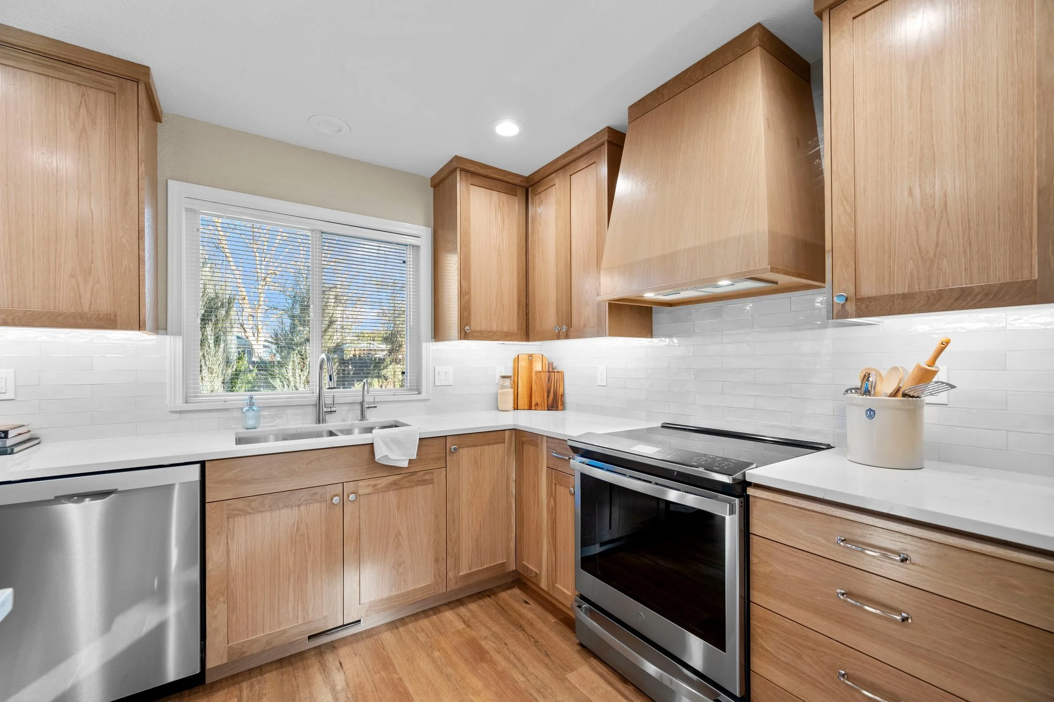 Kitchen with wooden cabinets, white countertops, stainless steel stove, and a window with blinds showing trees outside.
