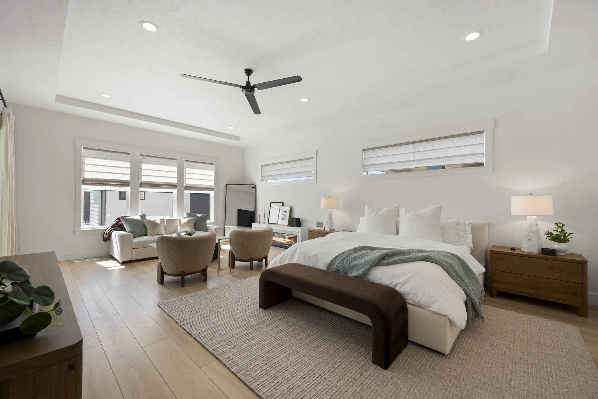 Bright bedroom with large bed, white bedding, gray blanket, wooden nightstand with lamp and plant, beige rug, white walls, multiple windows with blinds, ceiling fan, and a sitting area with armchairs and a sofa near a fireplace.