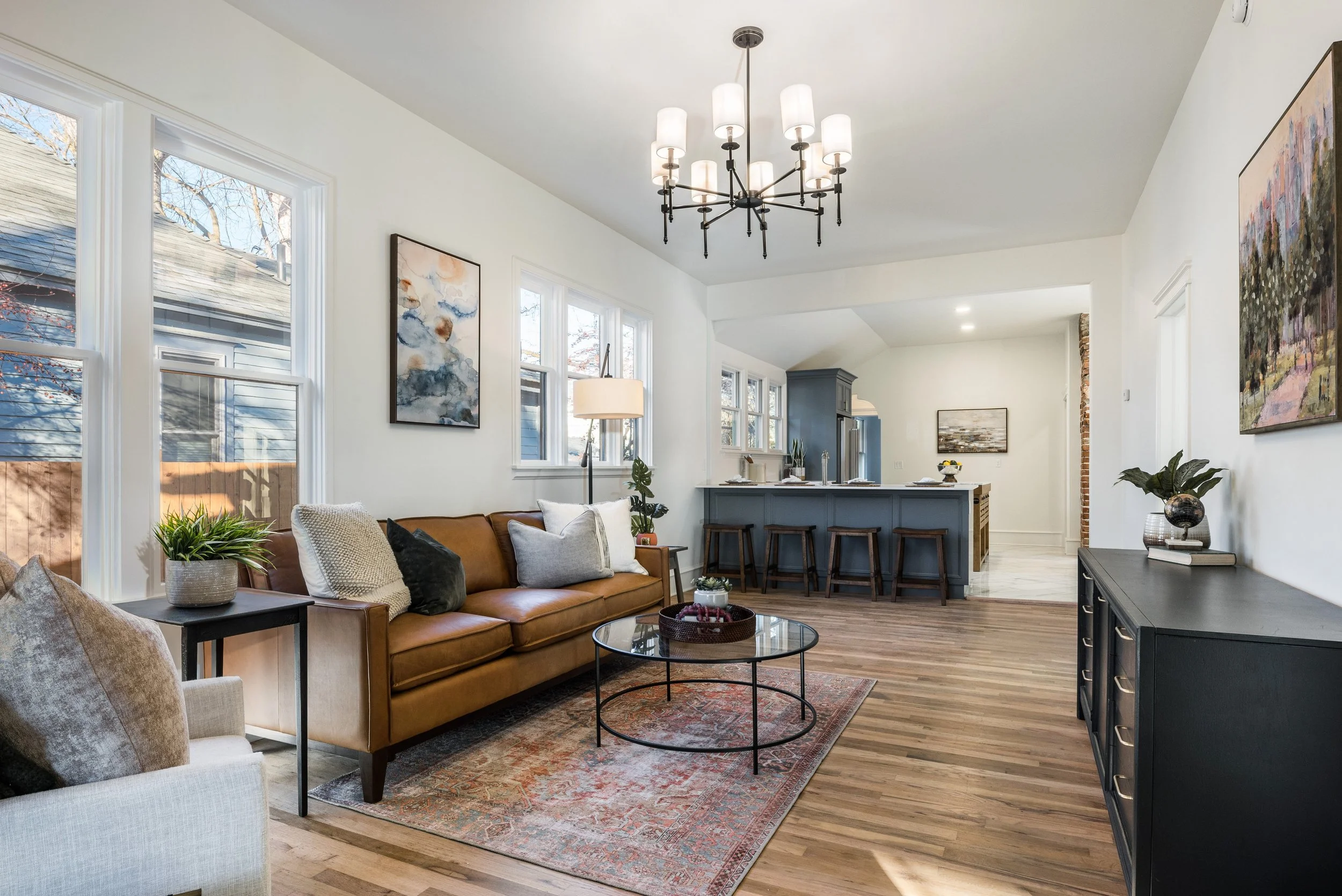 Living room featuring large windows, a brown leather sofa with pillows, a round black coffee table, an off-white armchair, and artwork on white walls. There is a modern chandelier hanging from the ceiling, a dark blue kitchen island with barstools, and a dark sideboard with decorative items. The room has wooden flooring and a mix of contemporary and cozy decor.