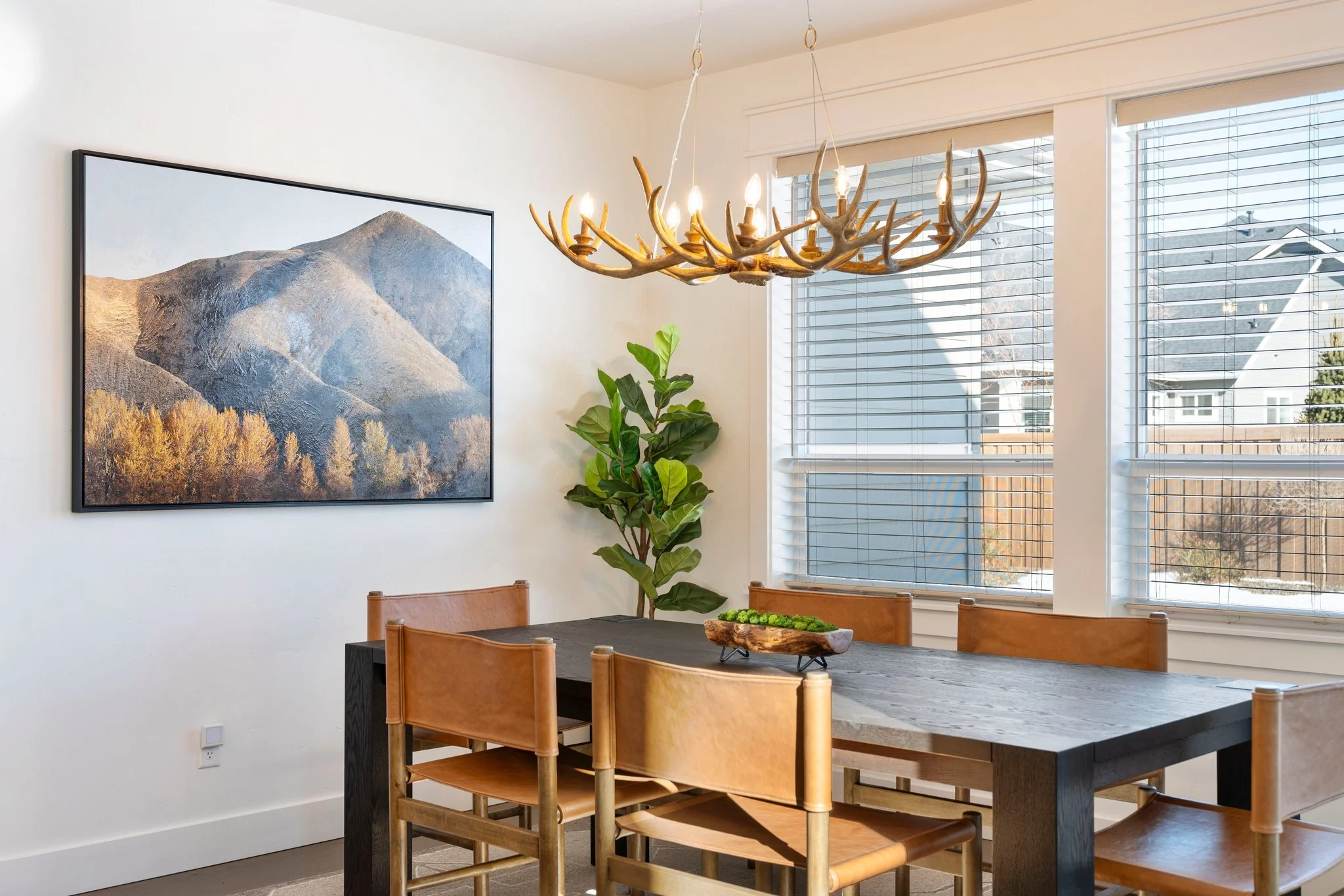 A dining room with a dark wooden table, four wooden chairs with tan leather seats, a large window with white blinds, a green potted plant, and artwork of mountain scenery on the wall. A chandelier made of antlers hangs above the table.