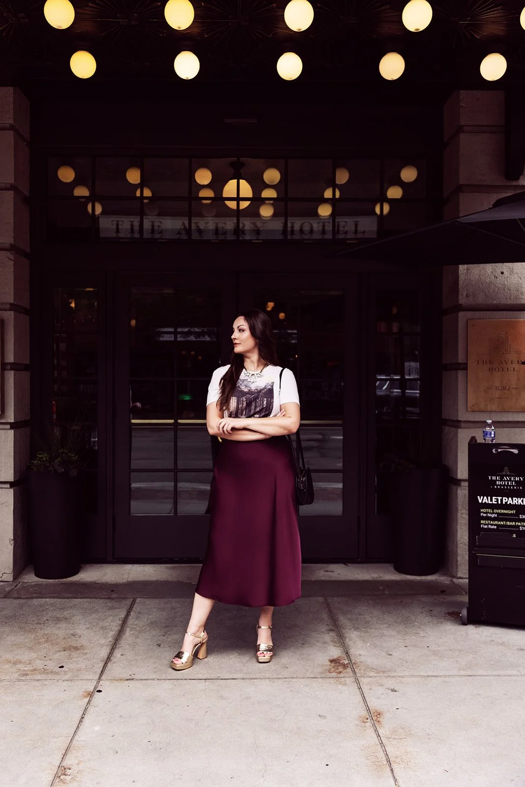 A woman standing outside the entrance of The Avery Hotel, dressed in a white t-shirt with a graphic, a maroon skirt, and high heels, with her arms crossed, in front of glass doors with warm lighting inside and a sign indicating valet parking.