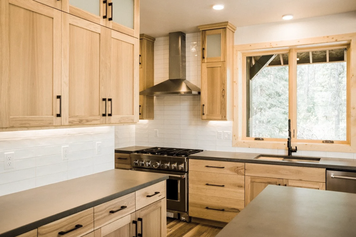 A kitchen with light wood cabinets, a stainless steel stove with oven, a black faucet over a sink, and a large window showing greenery outside.