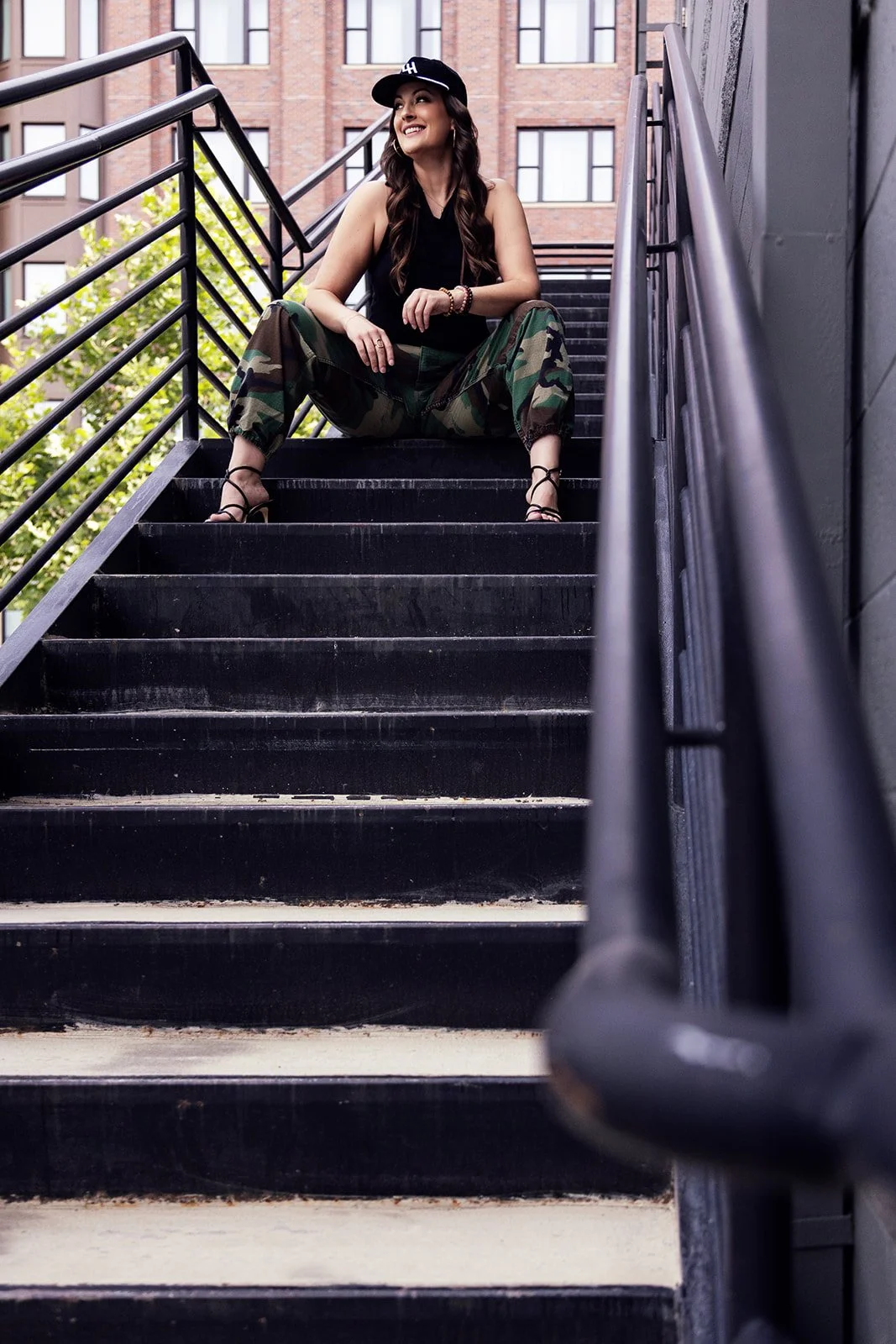 A woman sitting on outdoor black stairs with metal railings, smiling and looking to the side, wearing a black hat, black top, camouflage pants, and black sandals, with a background of a brick building and trees.