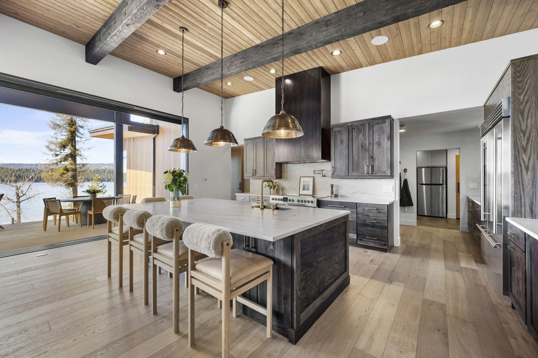 Modern kitchen with wooden ceiling beams, pendant lights, white cabinetry, and views of a lake through large windows.
