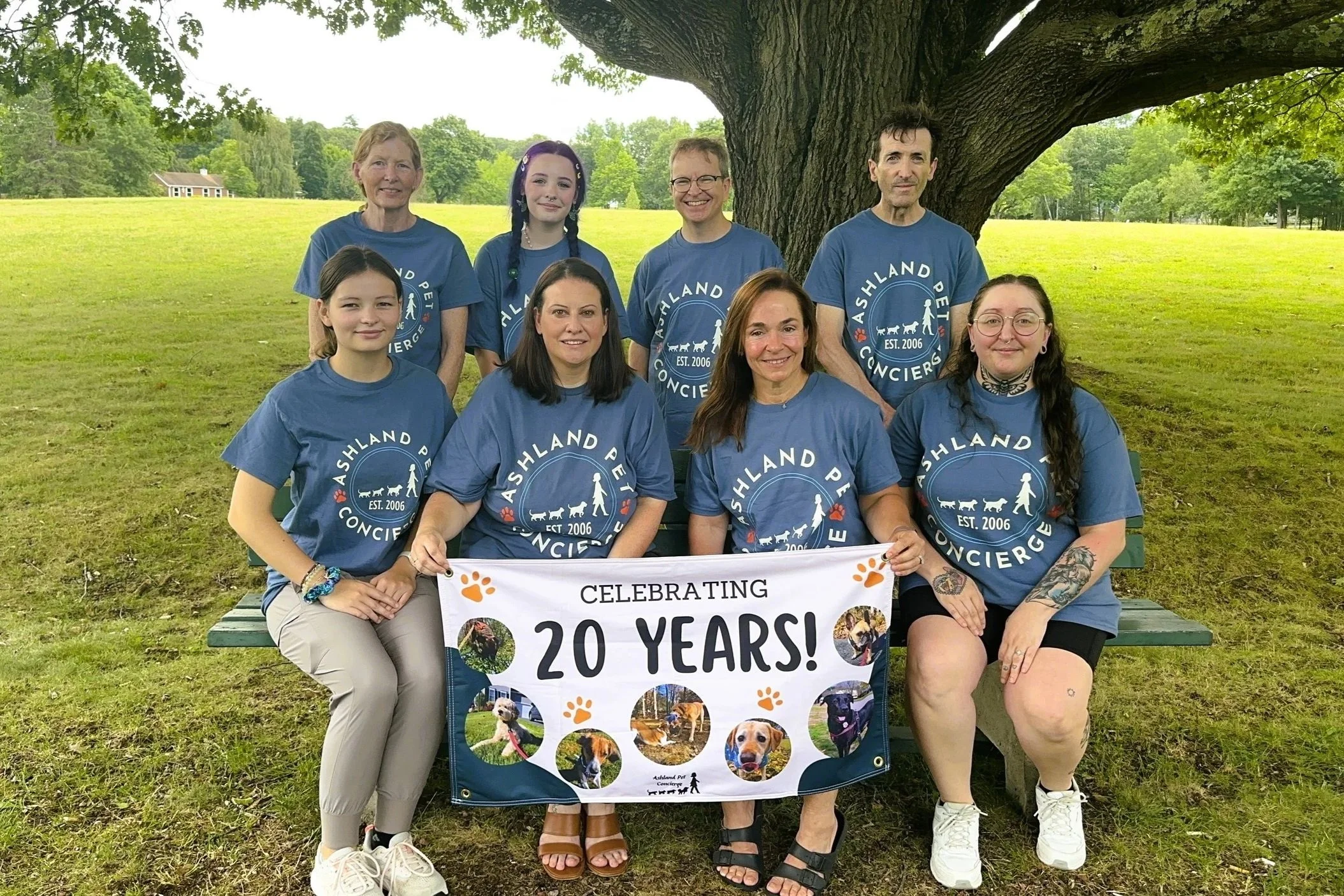 Group of nine people, including children and adults, posing outdoors under a large tree, holding a banner celebrating 20 years of an organization called Ashland Pet Concierges, all wearing matching blue T-shirts with the organization's logo, with a grassy field and small house in the background.