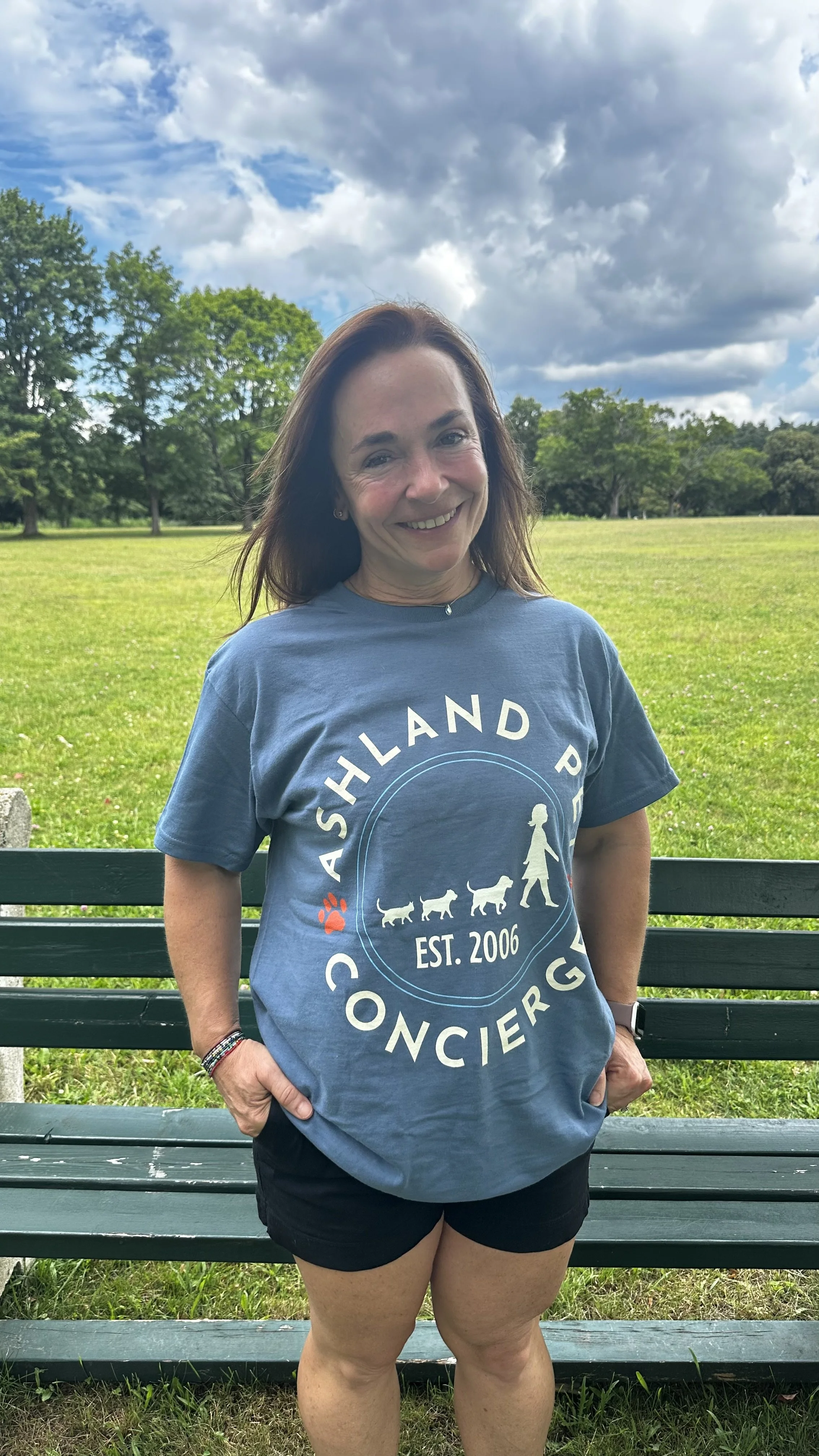 A woman smiling in a park, standing in front of a green bench with open grassy field and trees behind her. She is wearing a blue T-shirt with white text and black shorts, with her hands in her pockets.