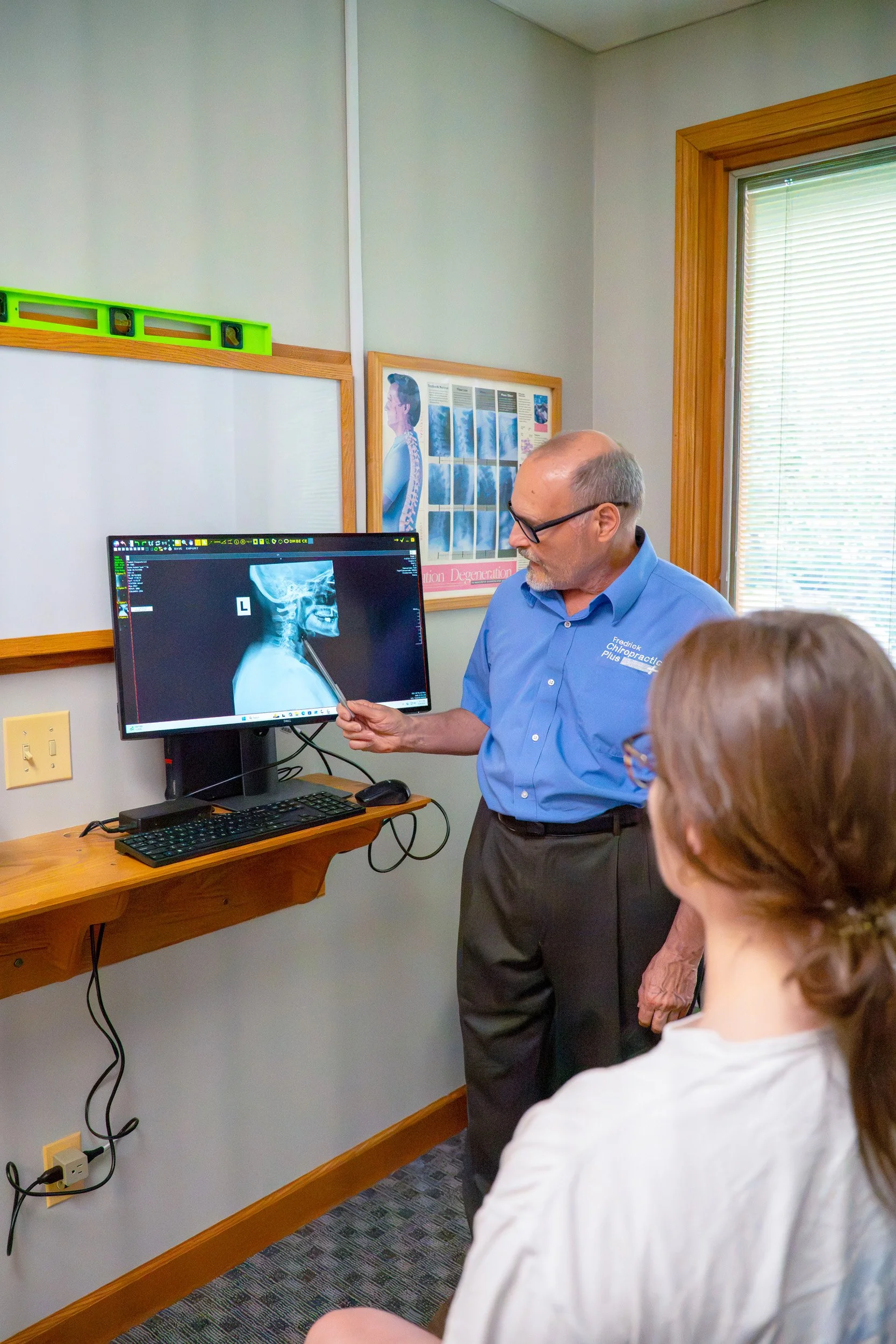 A chiropractor in a blue shirt teaches a group of people about a cervical spine X-ray, with a woman in white in the foreground and a woman with glasses in the background, in a room with a computer monitor, framed poster, and window.