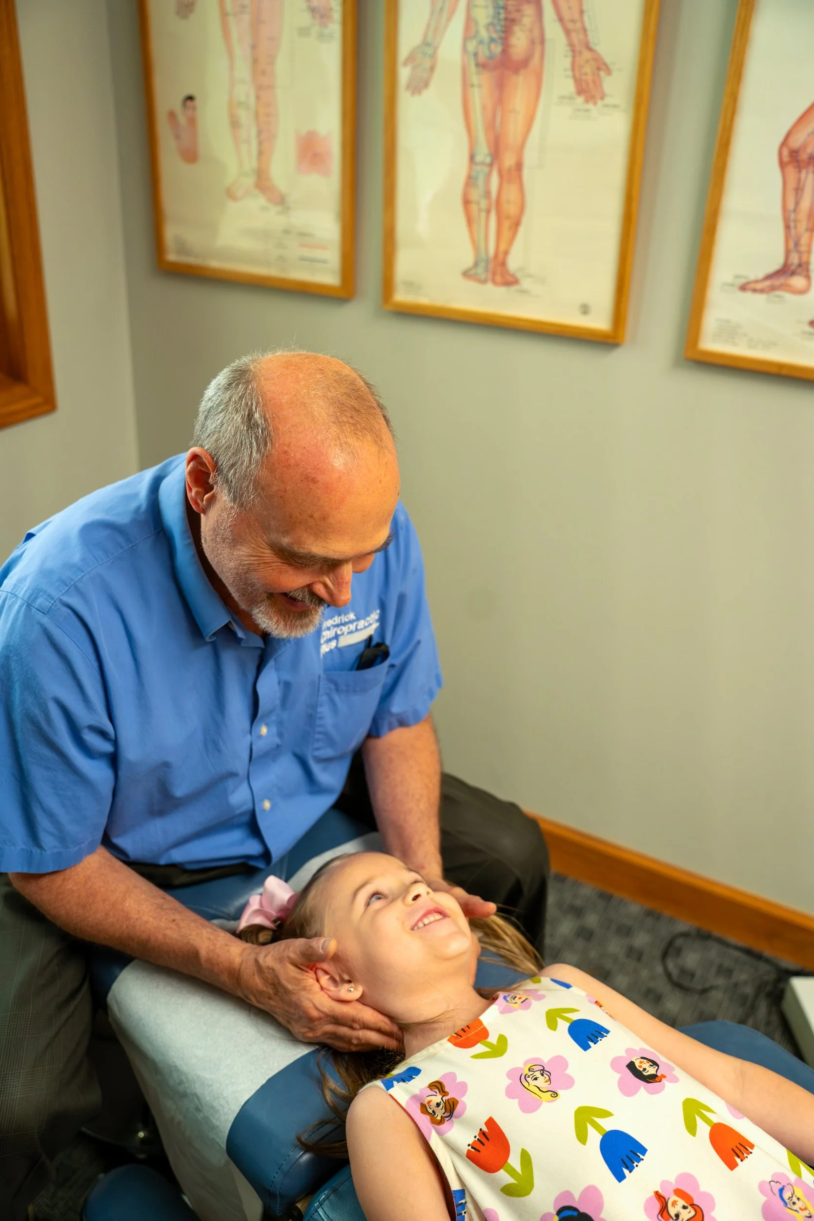 A young girl with long hair and a colorful dress lying on a chiropractic table, smiling up at a man with a beard and glasses, wearing a blue uniform, who is adjusting her neck. Behind them are framed anatomical posters on the wall.