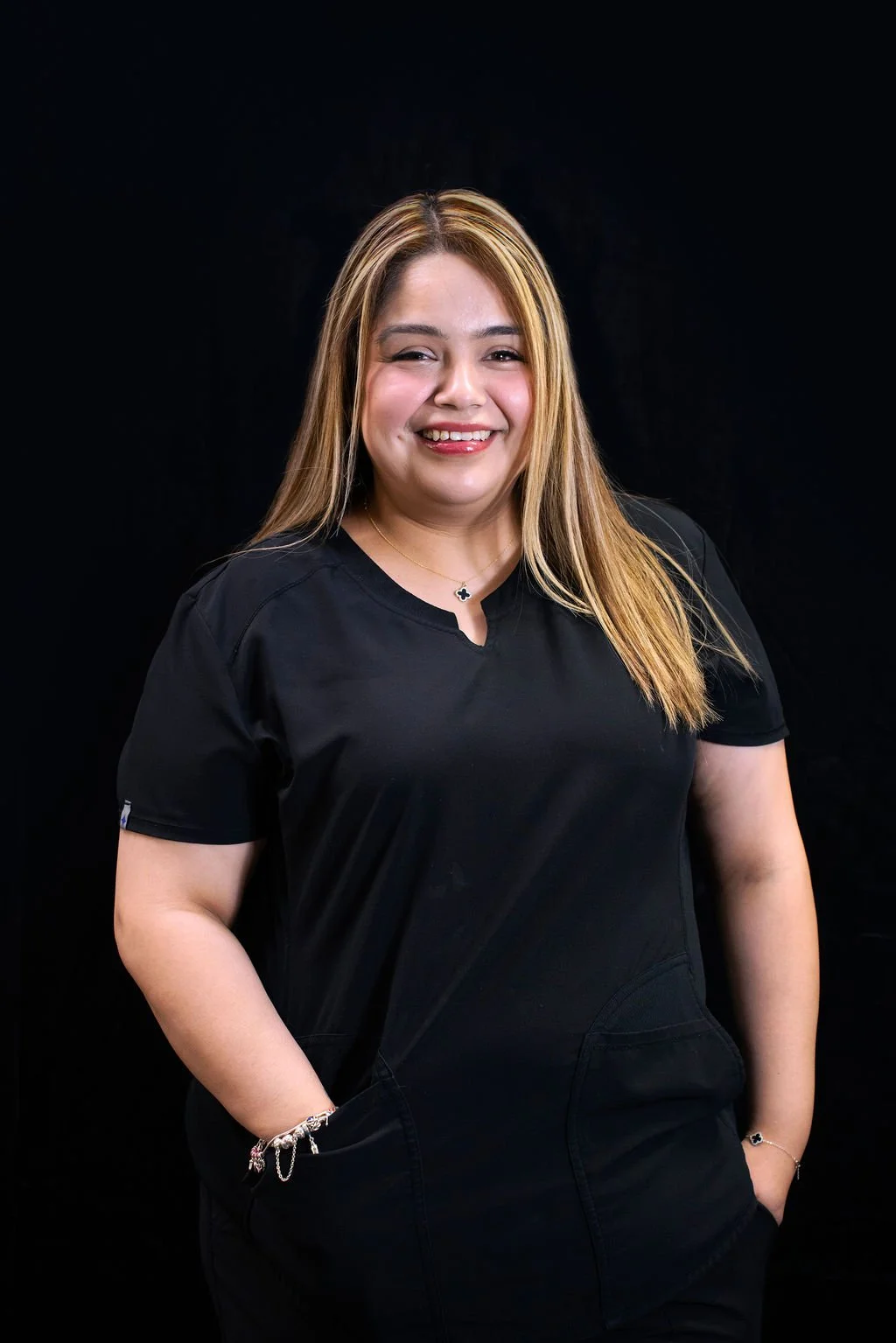 A woman with long blonde hair, wearing a black shirt, smiling and winking, standing against a black background.