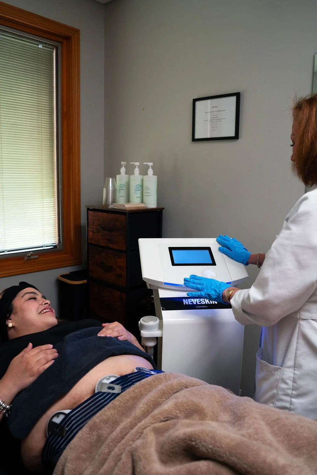 A woman lying on a medical bed, smiling and receiving a cosmetic treatment from a healthcare professional wearing blue gloves in a clinic room.