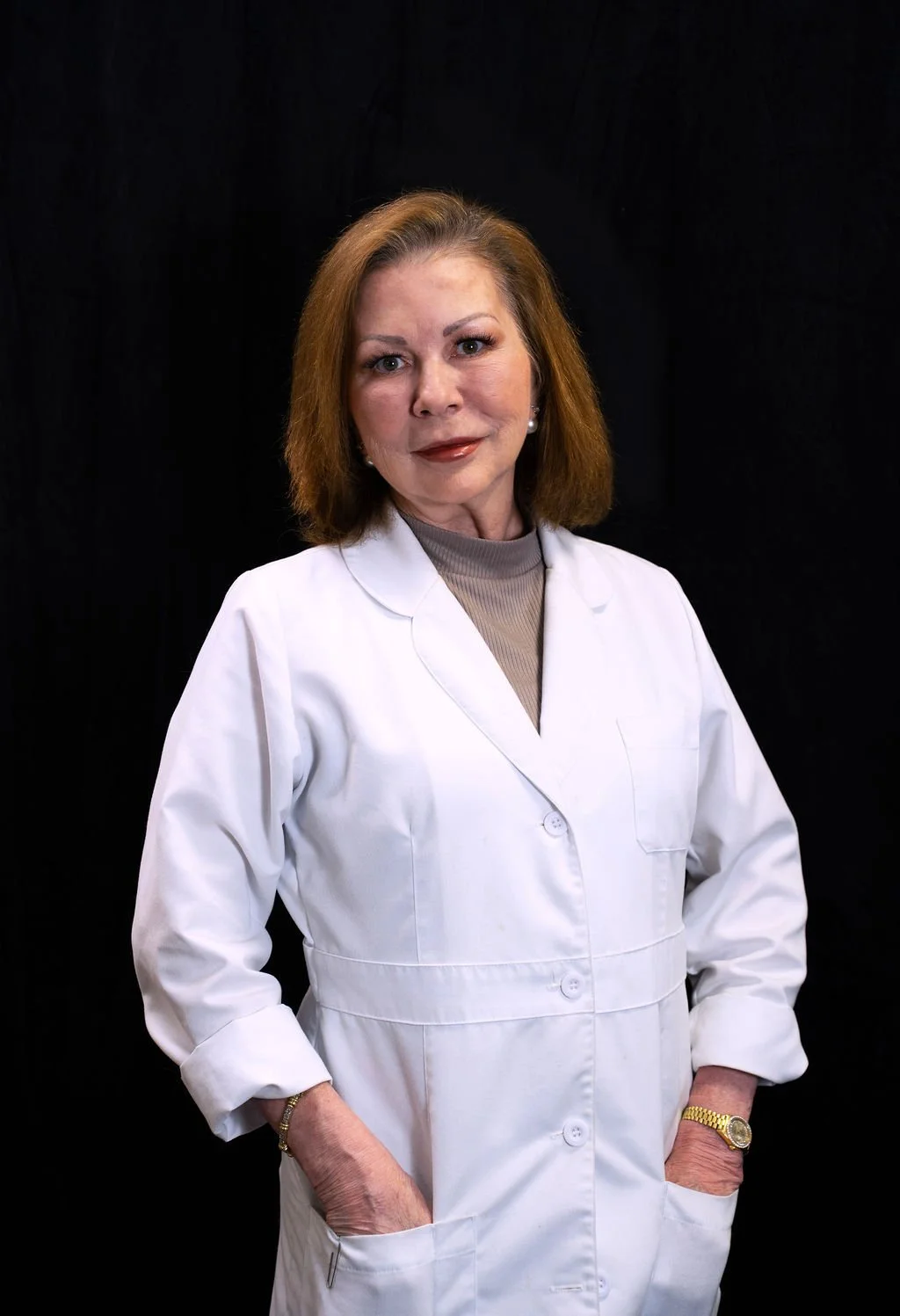 A woman in a white medical coat standing against a black background, with hands in pockets, looking at the camera.