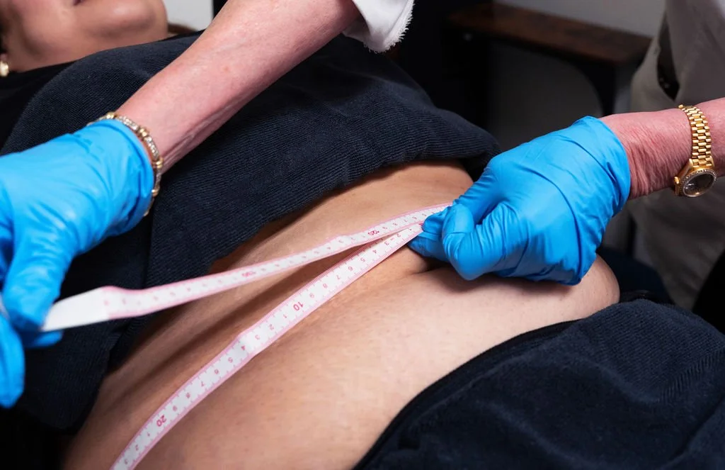A person wearing blue gloves measures a person's waist with a tape measure in a healthcare setting.