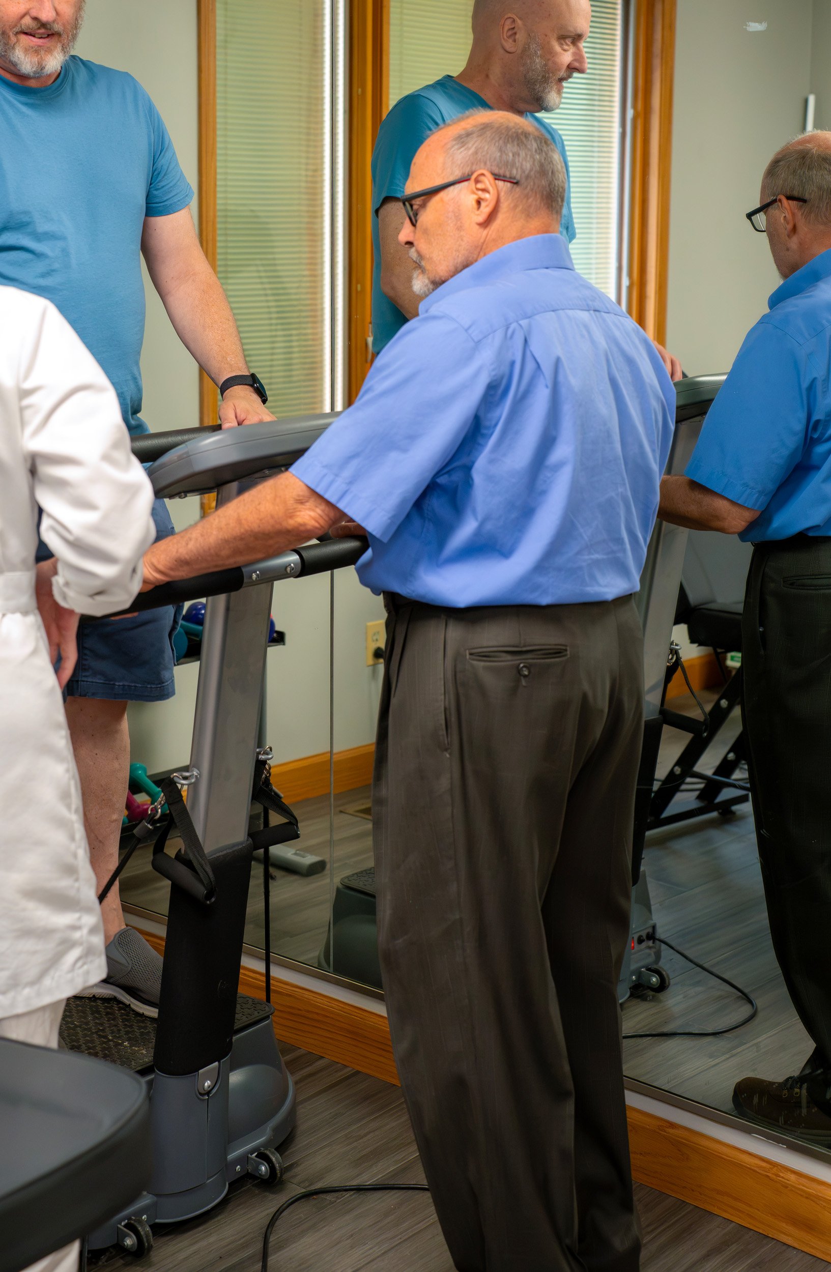 A group of middle-aged men are exercising on treadmills in a gym, wearing blue shirts and glasses, with wood-paneled walls and a large mirror.
