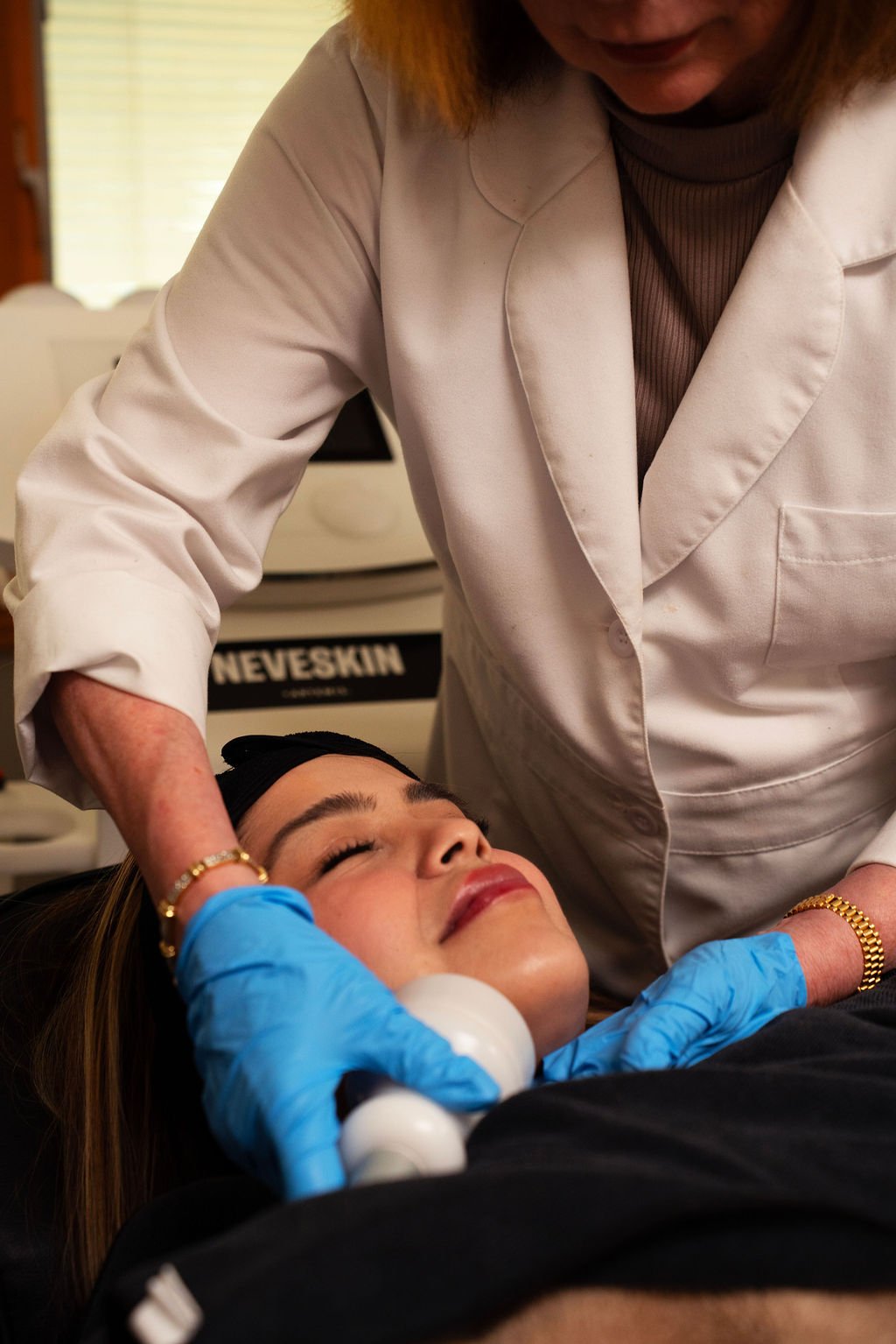 A cosmetologist performing a facial treatment on a woman lying down with her eyes closed in a spa or clinic setting.