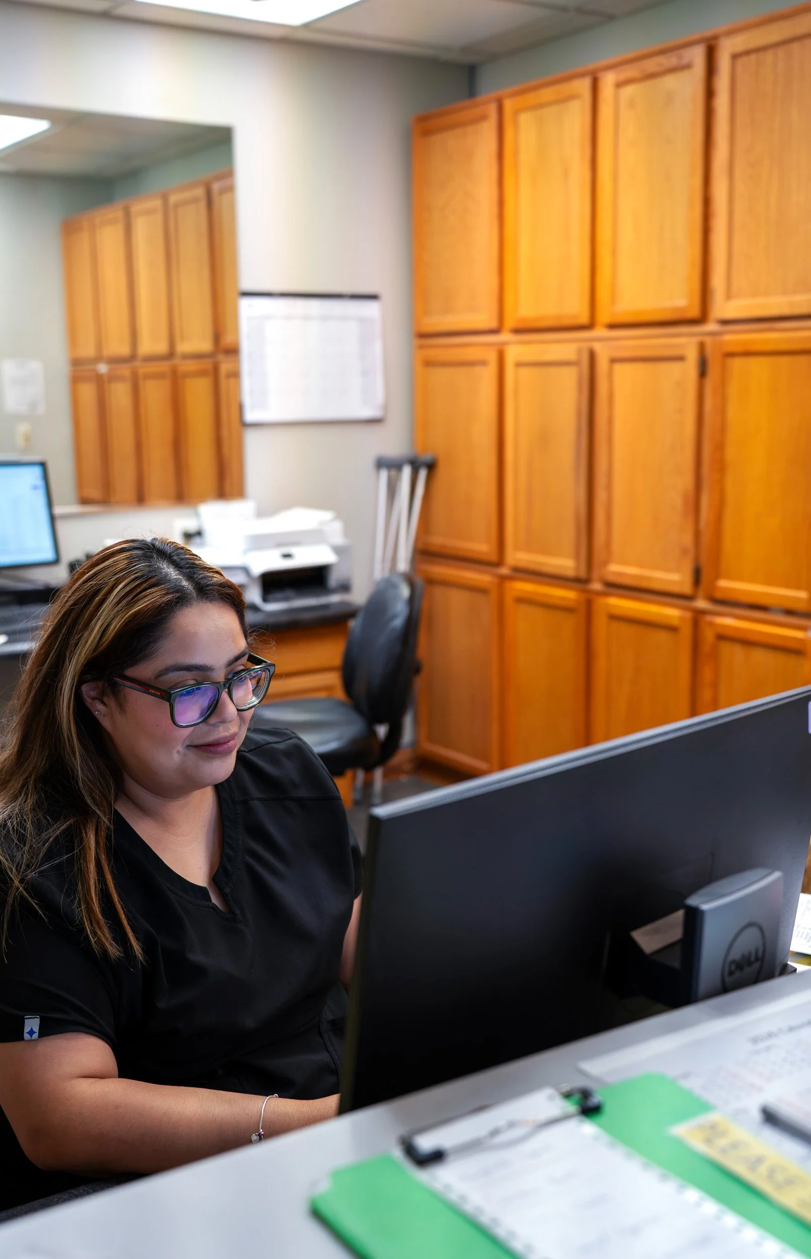 A woman with long brown hair, glasses, and a black shirt working at a computer in an office with wooden cabinets on the wall.