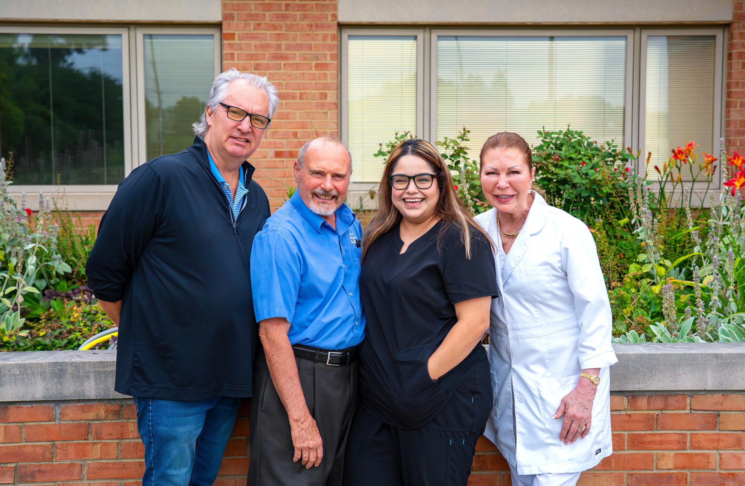 Four people standing outside in front of a brick building and garden, smiling for a photo. Two men on the left and woman on the right are dressed casually, while the woman in the middle wears scrubs, indicating she may be a healthcare worker.