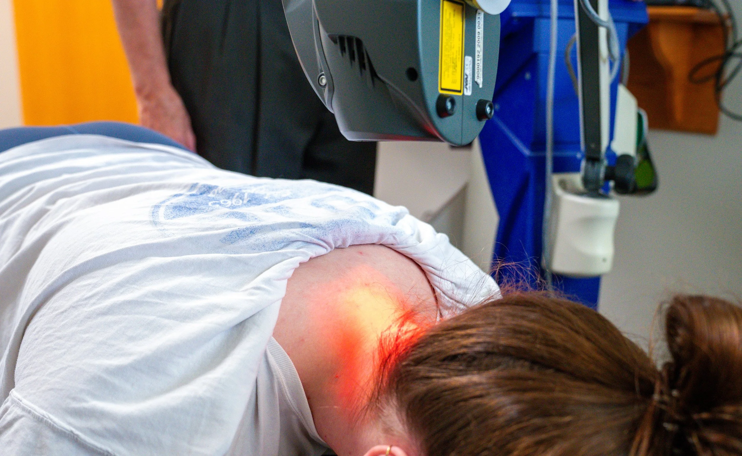 A person lying on an examination table receiving a laser or light therapy treatment on their shoulder or upper back area, with medical equipment in the background.