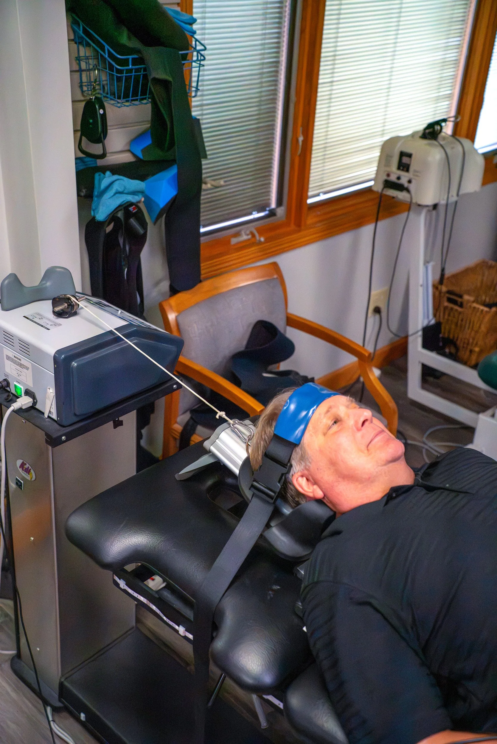 A man lying on a medical examination table with electrodes and a headband, connected to medical equipment in a room with wooden window frames and blinds, with various medical and personal items in the background.