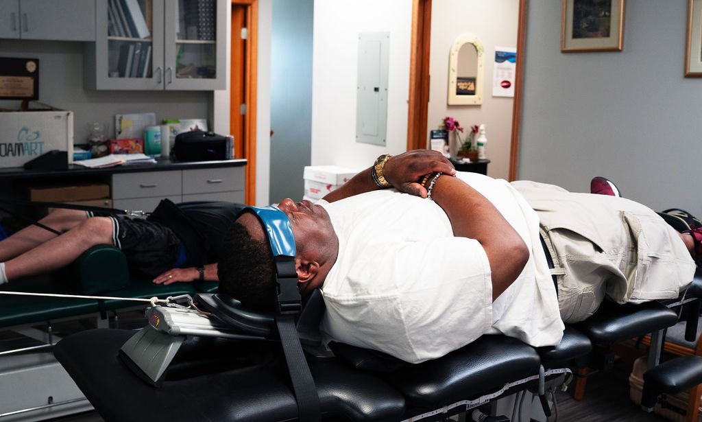Two people lying on medical examination tables in a room with medical equipment and decor. The man in the foreground is resting with hands clasped, wearing a white shirt and khaki pants, while the person in the background is a child lying face down with legs extended, wearing dark clothing.