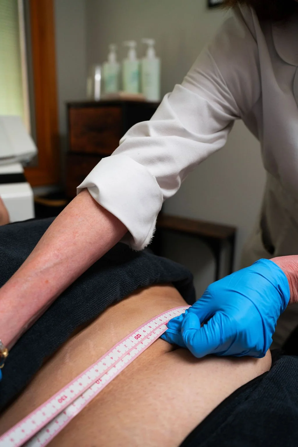A healthcare professional wearing blue gloves measures a patient's waist with a pink measuring tape.