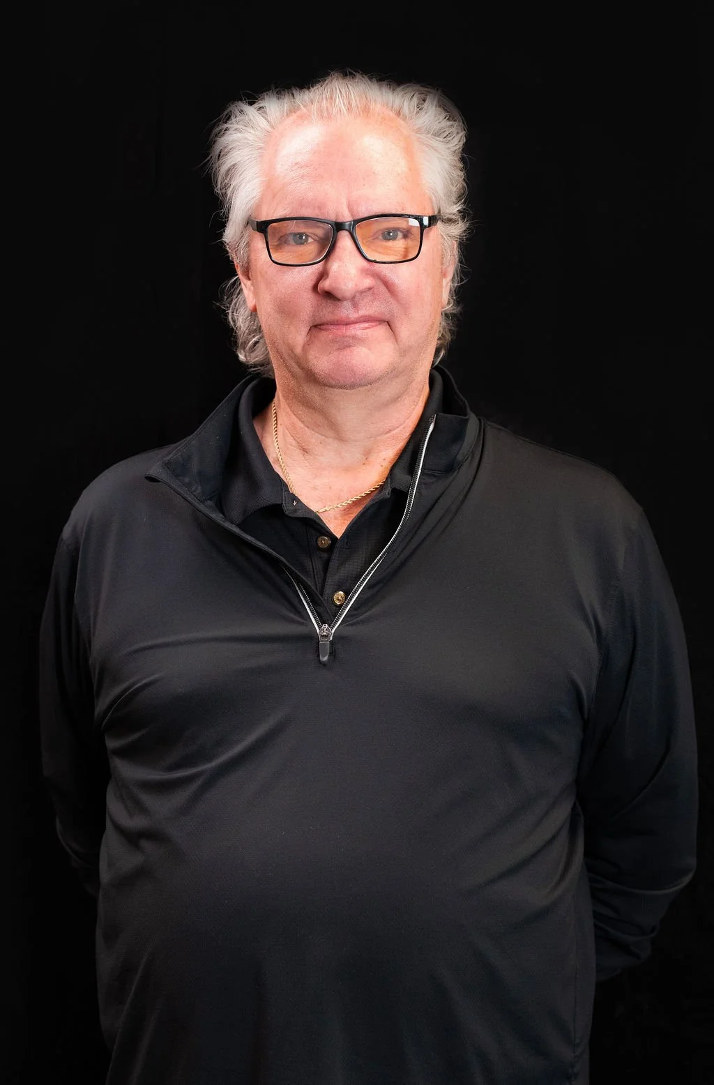 An older man with white hair, glasses, wearing a black jacket and gold chain, standing against a black background.