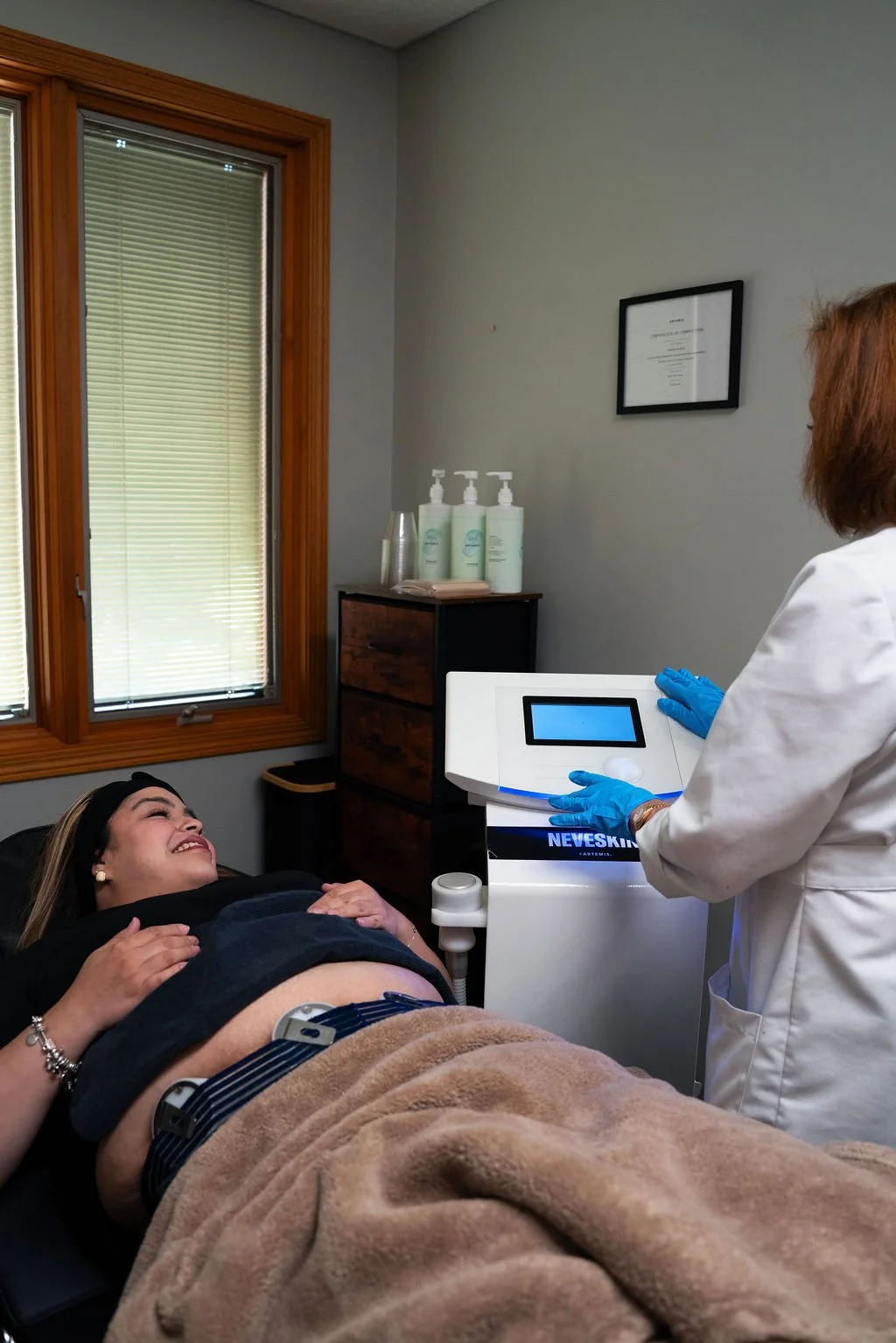 A woman lying on a medical examination table smiling while a healthcare professional in a white coat and blue gloves operates a medical device in a room with a window, wood trim, and bottles of lotion on a small cabinet.