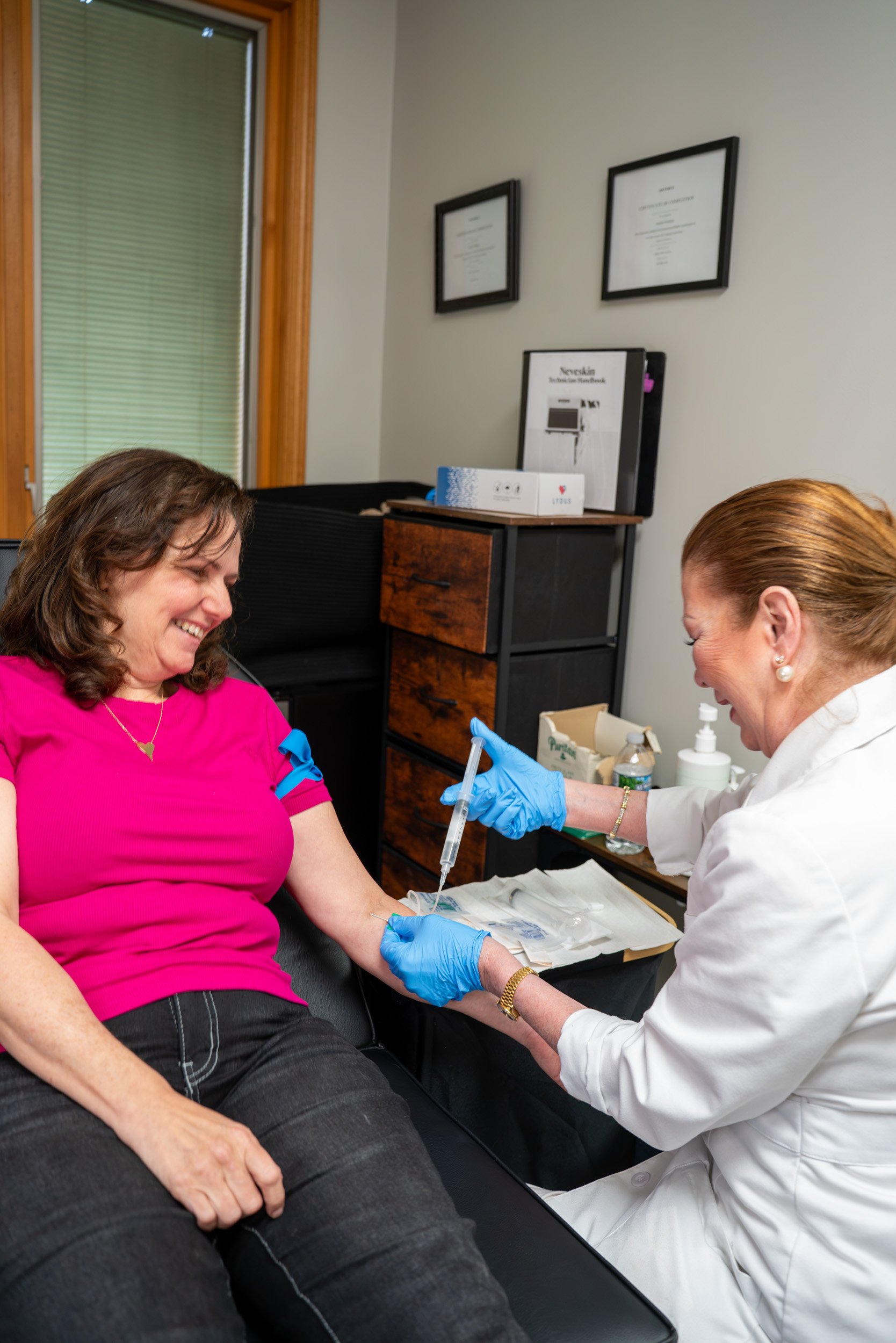A woman in a pink shirt sitting in a chair while healthcare worker in white coat and blue gloves draws blood from her arm with a syringe. The woman is smiling and there are framed certificates on the wall.