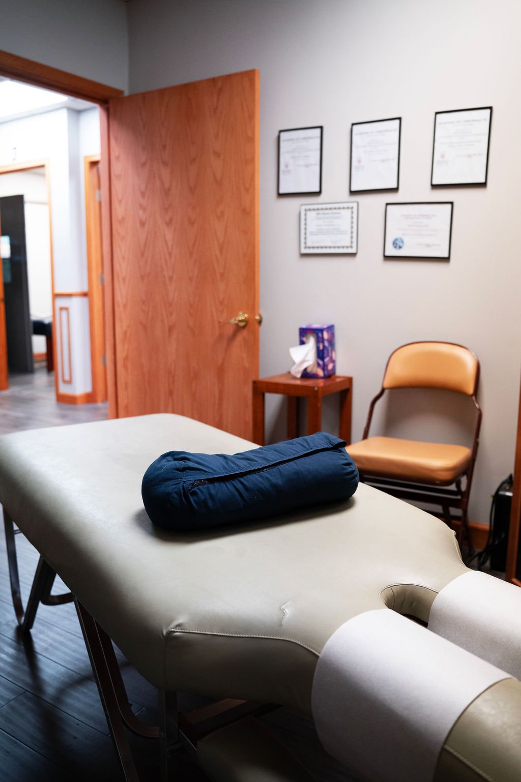 A medical examination room with a beige exam table, a blue pillow, a small wooden side table with tissues and a box of tissues, a beige chair, and wall certificates. An open wooden door leads to another room.
