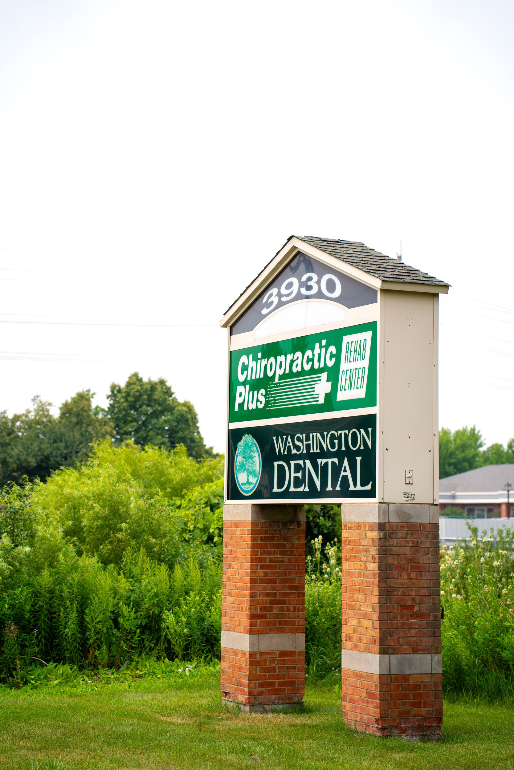 Sign for Chiropractic Plus and Washington Dental, with a brick base and a gray roof, standing in a green landscape with bushes and trees in the background.