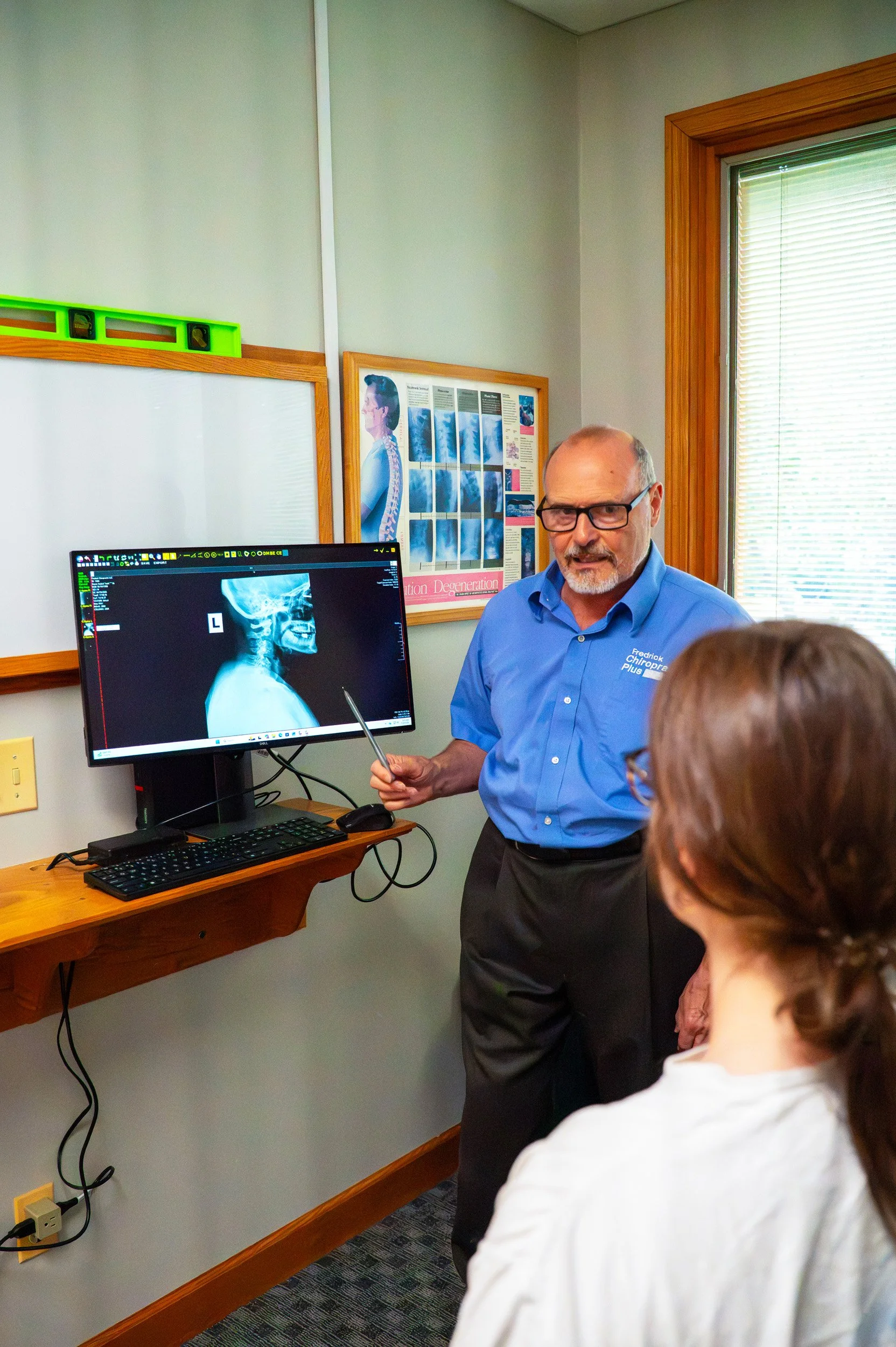 A man in a blue shirt demonstrates a neck X-ray to a group of people in a medical office, with a computer monitor displaying the X-ray and a safety level on the wall.
