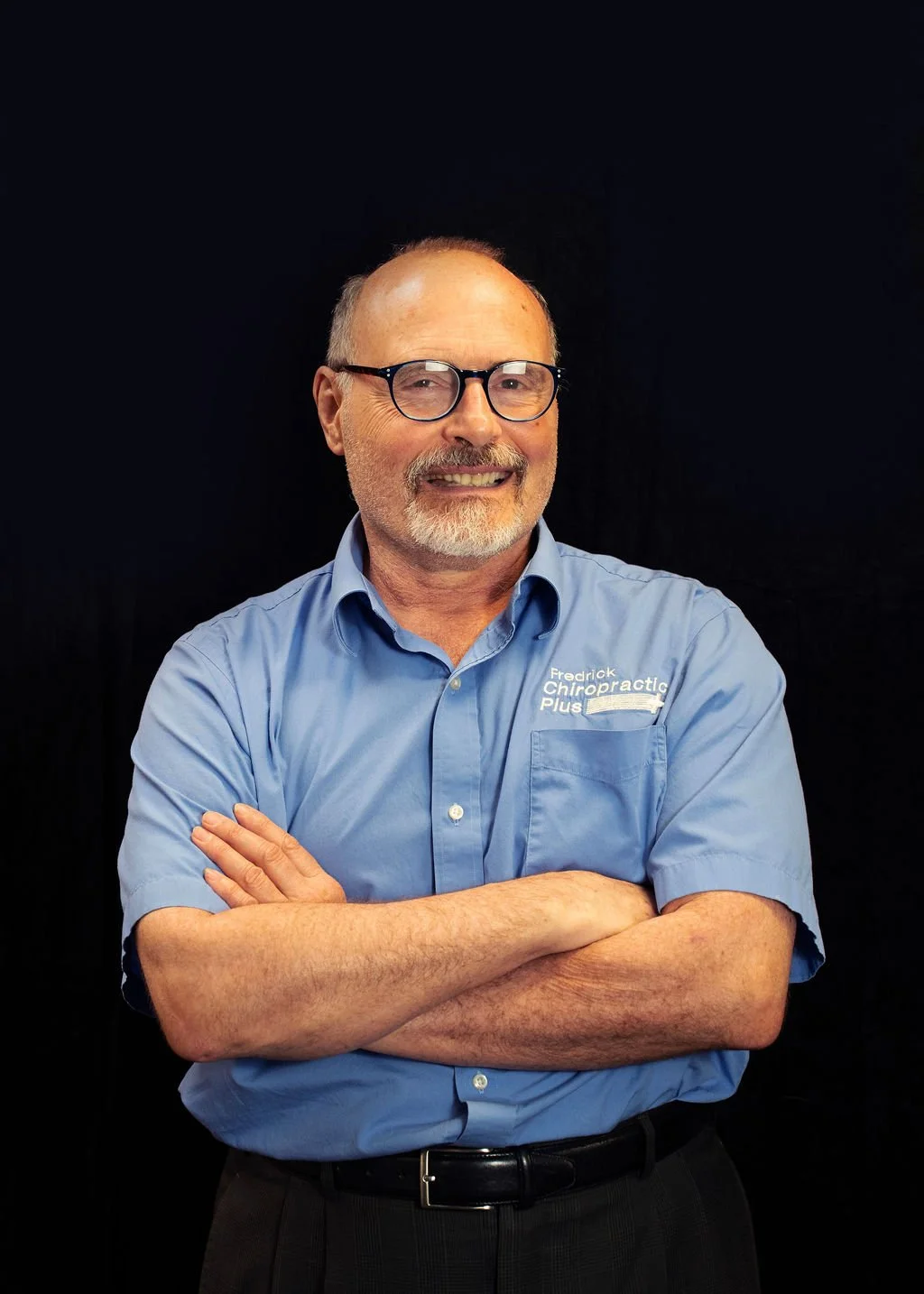 A man with glasses, a beard, and a mustache wearing a light blue shirt with 'Fredrick Chiropractic Plus' embroidery, smiling with arms crossed against a black background.