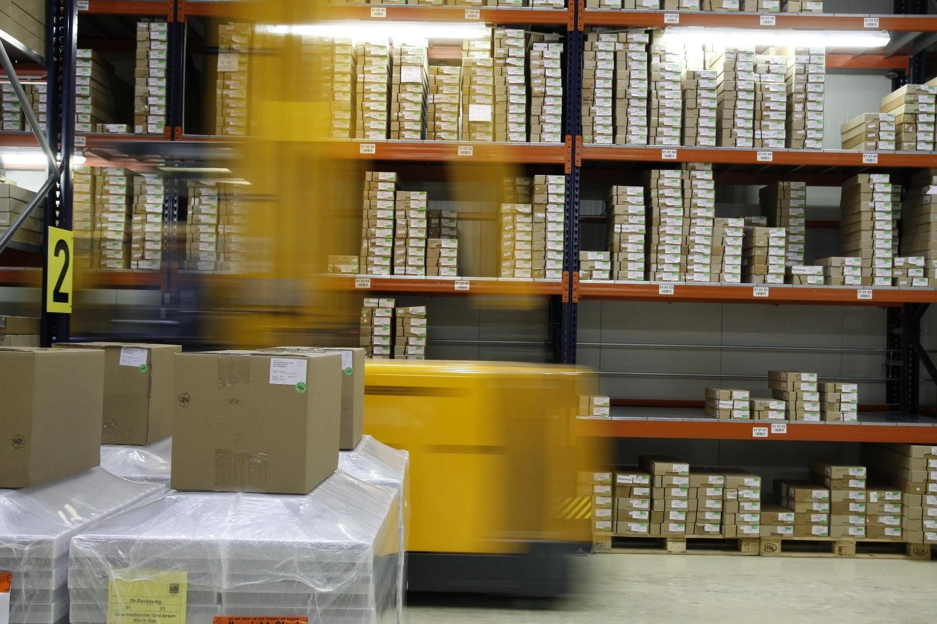 Warehouse with stacked cardboard boxes on orange and blue metal shelves and large wrapped boxed items on the floor.