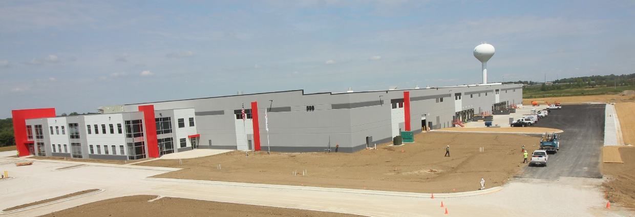 An industrial building under construction with a gray and red exterior, a large parking lot, and a water tower in the background.