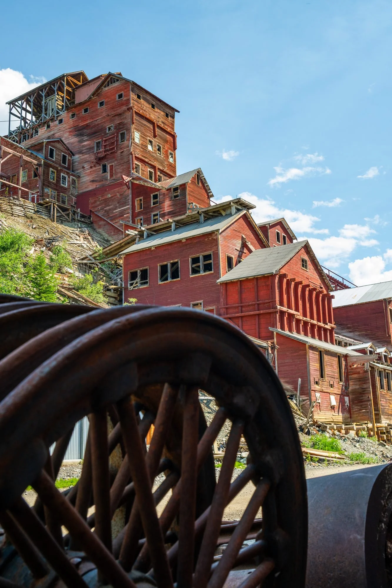 A view of a large, multi-story red wooden building with a complex structure, seen through a metal coiled object in the foreground. The building appears weathered, with some windows and parts under construction or in disrepair, under a blue sky with s
