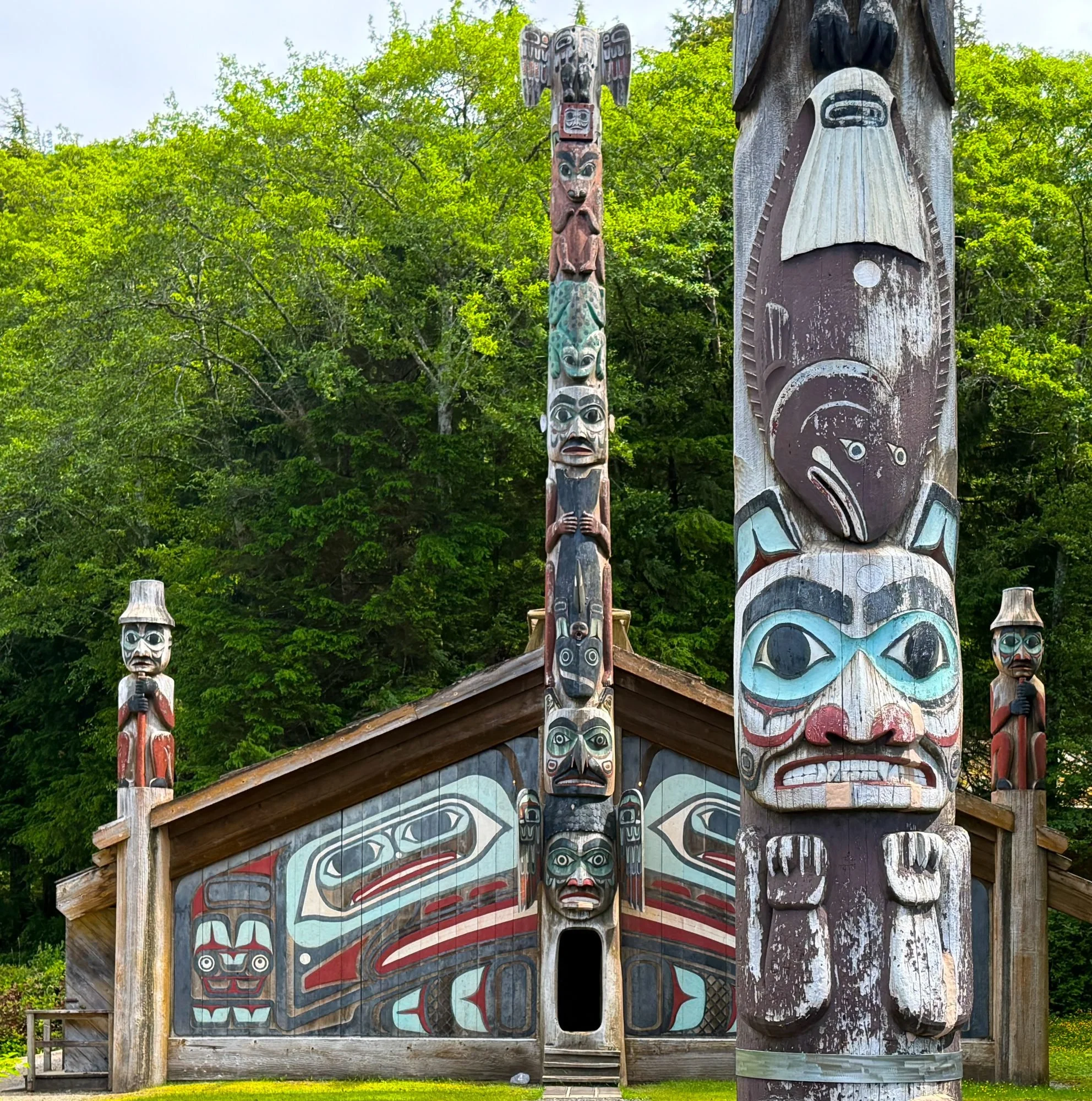 Colorful totem poles with traditional Indigenous artwork and faces, surrounded by green trees, part of a cultural display or historical site.