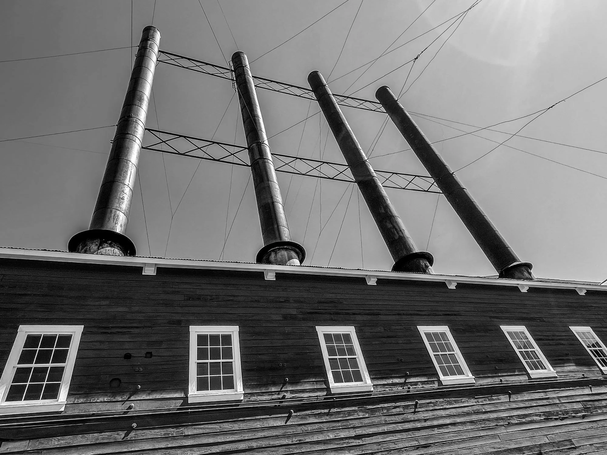 Black and white photo of a house roof with multiple windows and four tall, metal power line towers with multiple wires extending from them to the sky.