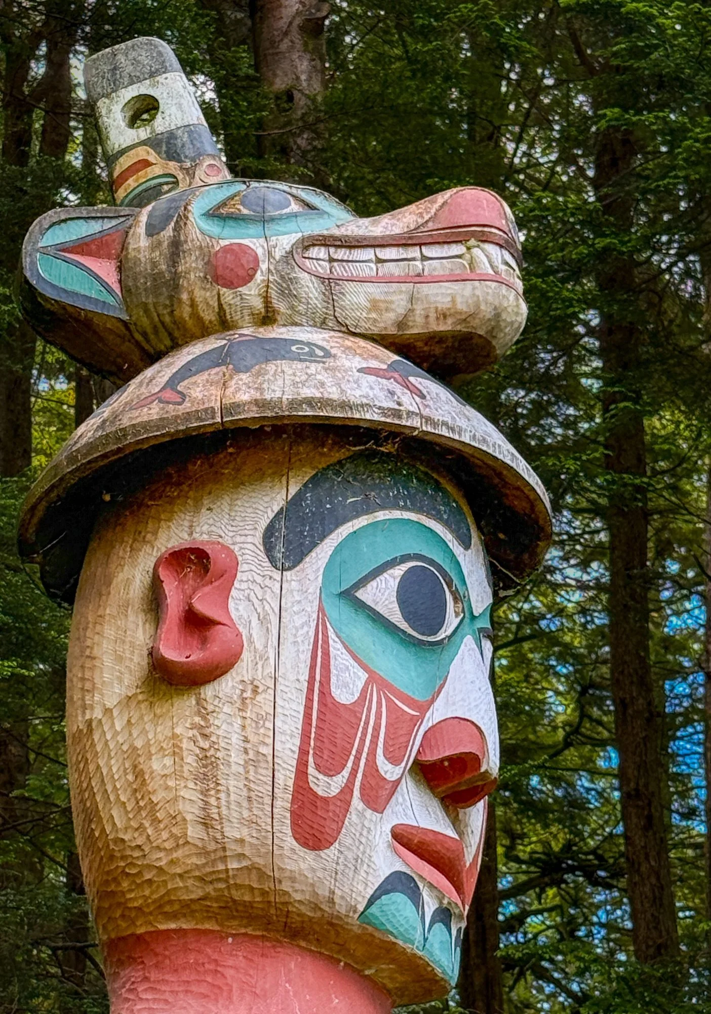 Colorfully painted totem pole in a forest setting with green trees in the background.