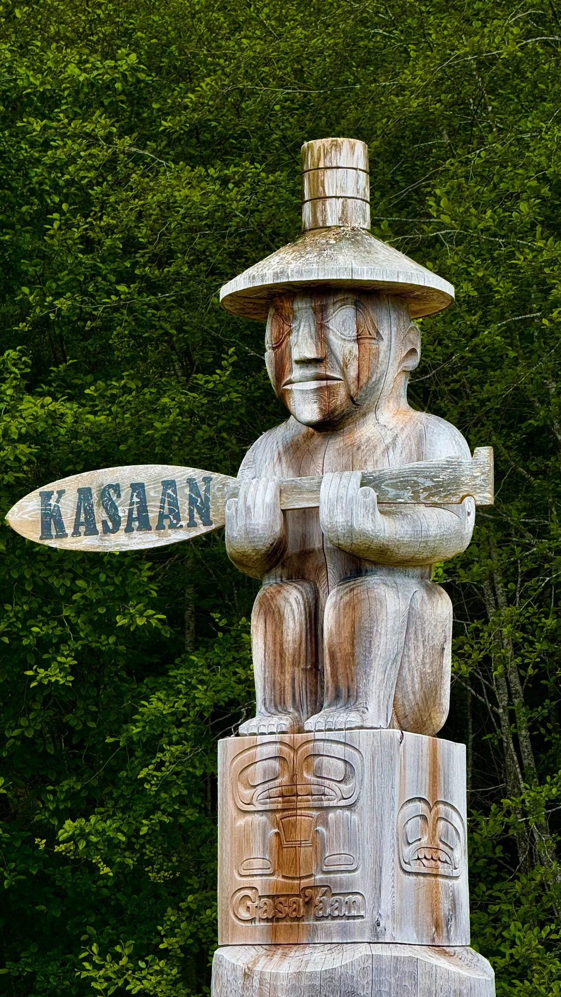 Wooden carved totem pole depicting a human figure wearing a wide hat and holding a paddle, with the word 'KASAN' on a sign on the paddle. The pole is displayed outdoors against green foliage.