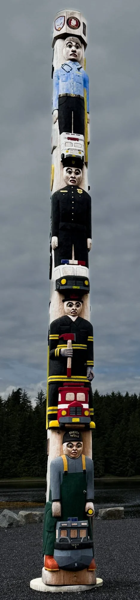 A tall totem pole featuring carved figures representing emergency personnel, including firefighters, police officers, paramedics, and rescue workers, painted in bright colors.