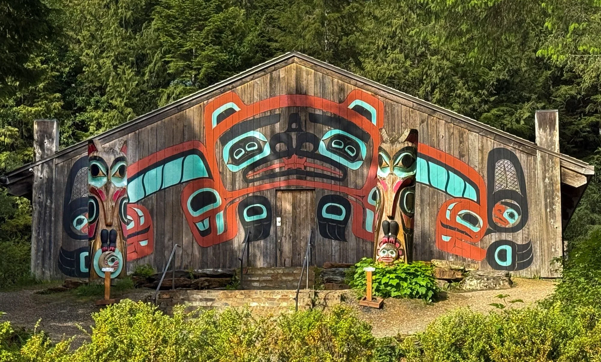A wooden building with colorful traditional Indigenous art and mask decorations on its facade, set against a background of green trees.