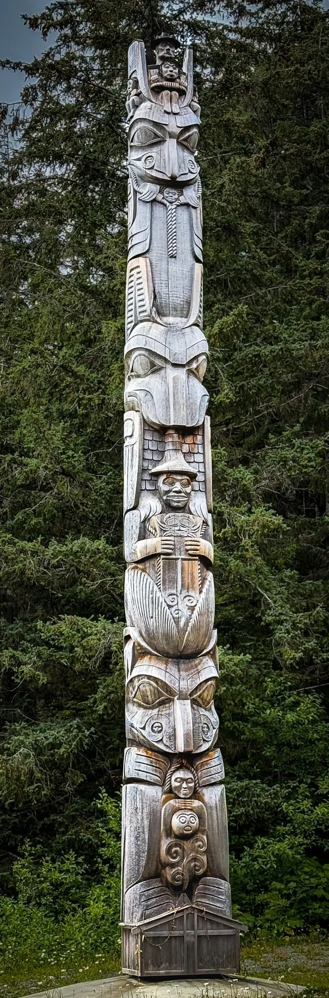 A tall wooden totem pole with multiple carved figures stacked vertically, set against a background of green trees.