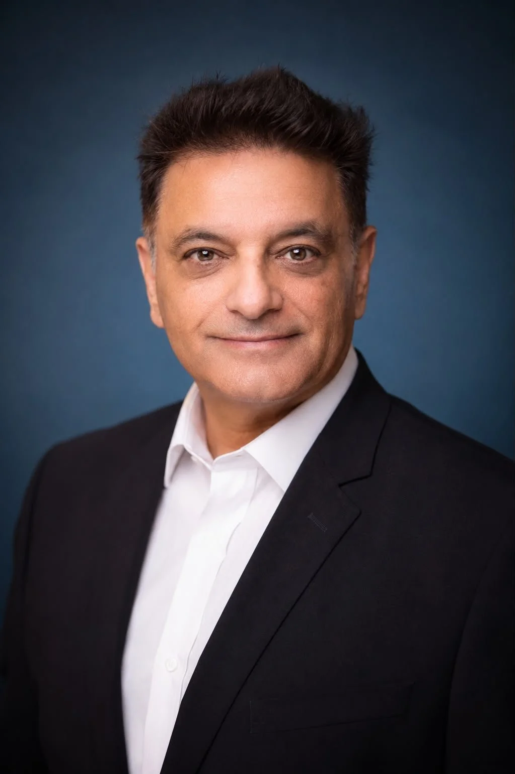 A professional headshot of Sanjay Kalra, a middle-aged man wearing a black suit and white shirt, smiling slightly, against a dark blue background.